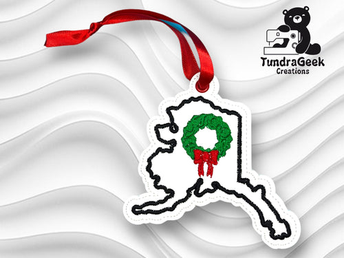 Embroidered ornament shaped like Alaska with a green wreath and red ribbon, displayed on a wavy white background with a TundraGeek Creations logo.