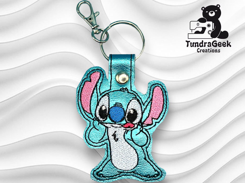 Embroidered blue cartoon character keychain with pink ears and a metal clip, displayed on a wavy white background. The logo TundraGeek Creations appears in the top right corner.