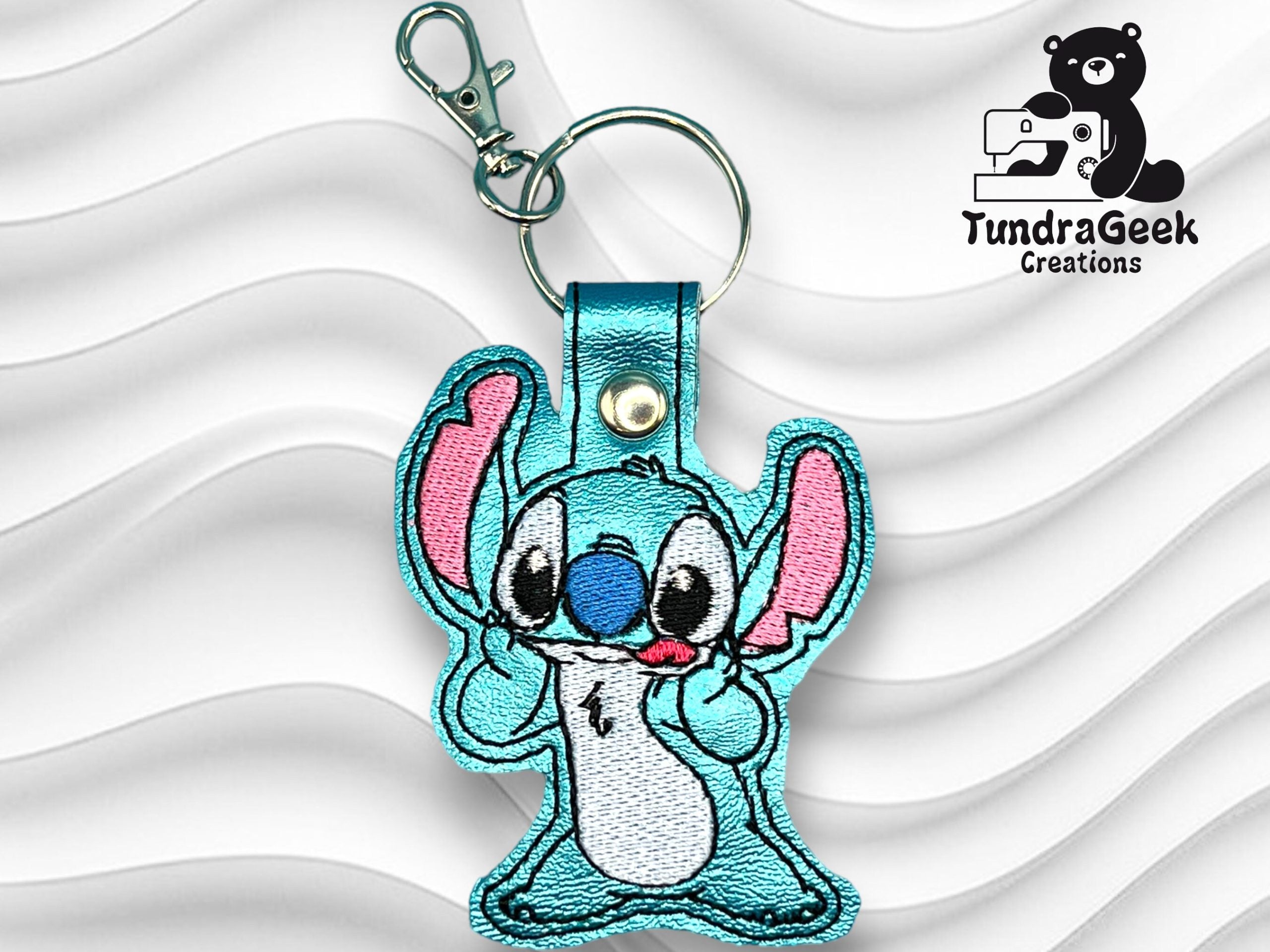 Embroidered blue cartoon character keychain with pink ears and a metal clip, displayed on a wavy white background. The logo TundraGeek Creations appears in the top right corner.