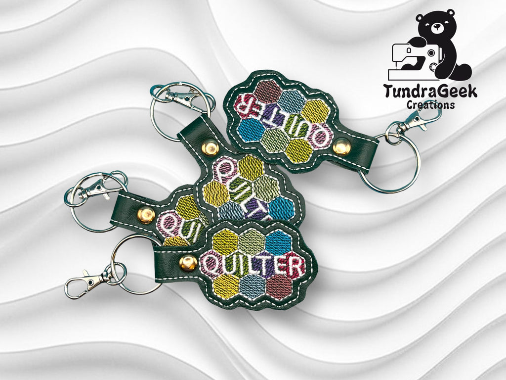 Three embroidered keychains with hexagon patterns and the text Hello Quilt Quilter, displayed on a wavy white background with a TundraGeek Creations logo featuring a bear.