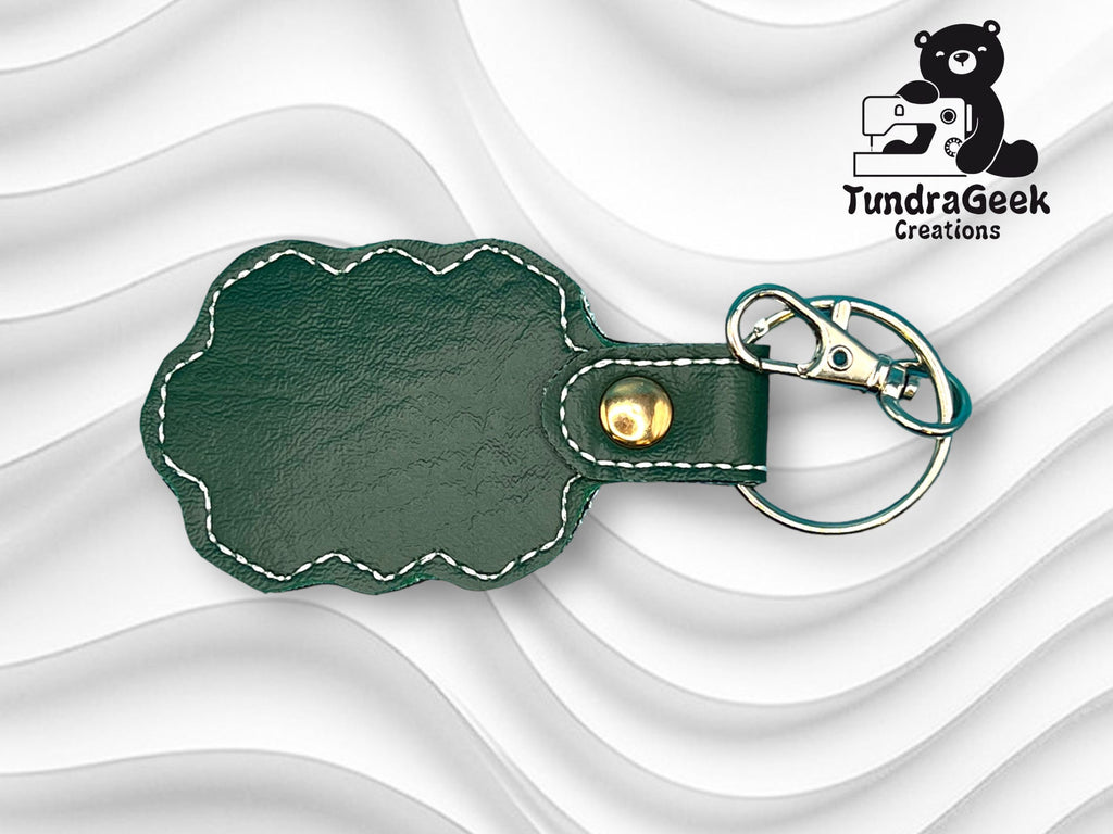 Green faux leather keychain with scalloped edges, white stitching, gold snap, and a metal keyring with clip. TundraGeek Creations logo with a bear and sewing machine in upper right corner.