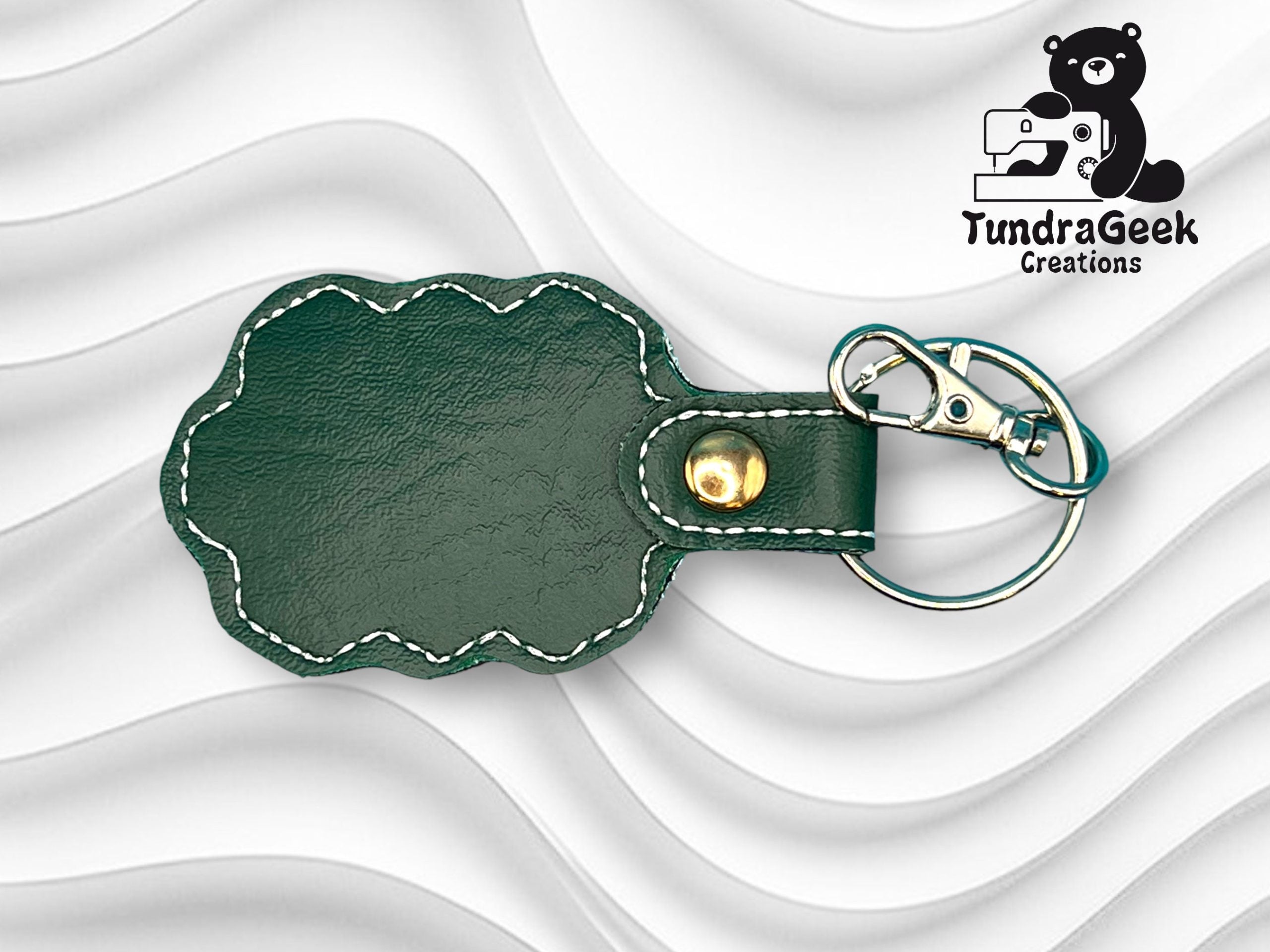 Green faux leather keychain with scalloped edges, white stitching, gold snap, and a metal keyring with clip. TundraGeek Creations logo with a bear and sewing machine in upper right corner.