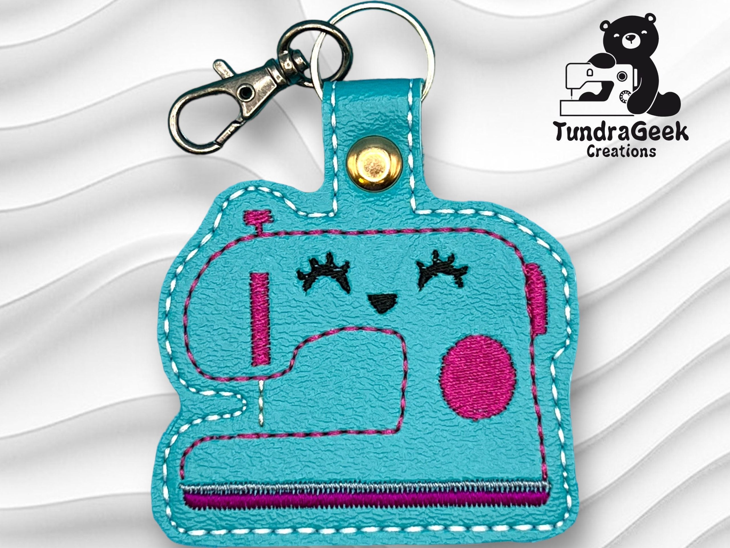 A blue faux leather keychain shaped like a sewing machine with pink details and stitched facial features, attached to a metal clasp; TundraGeek Creations logo in the corner.