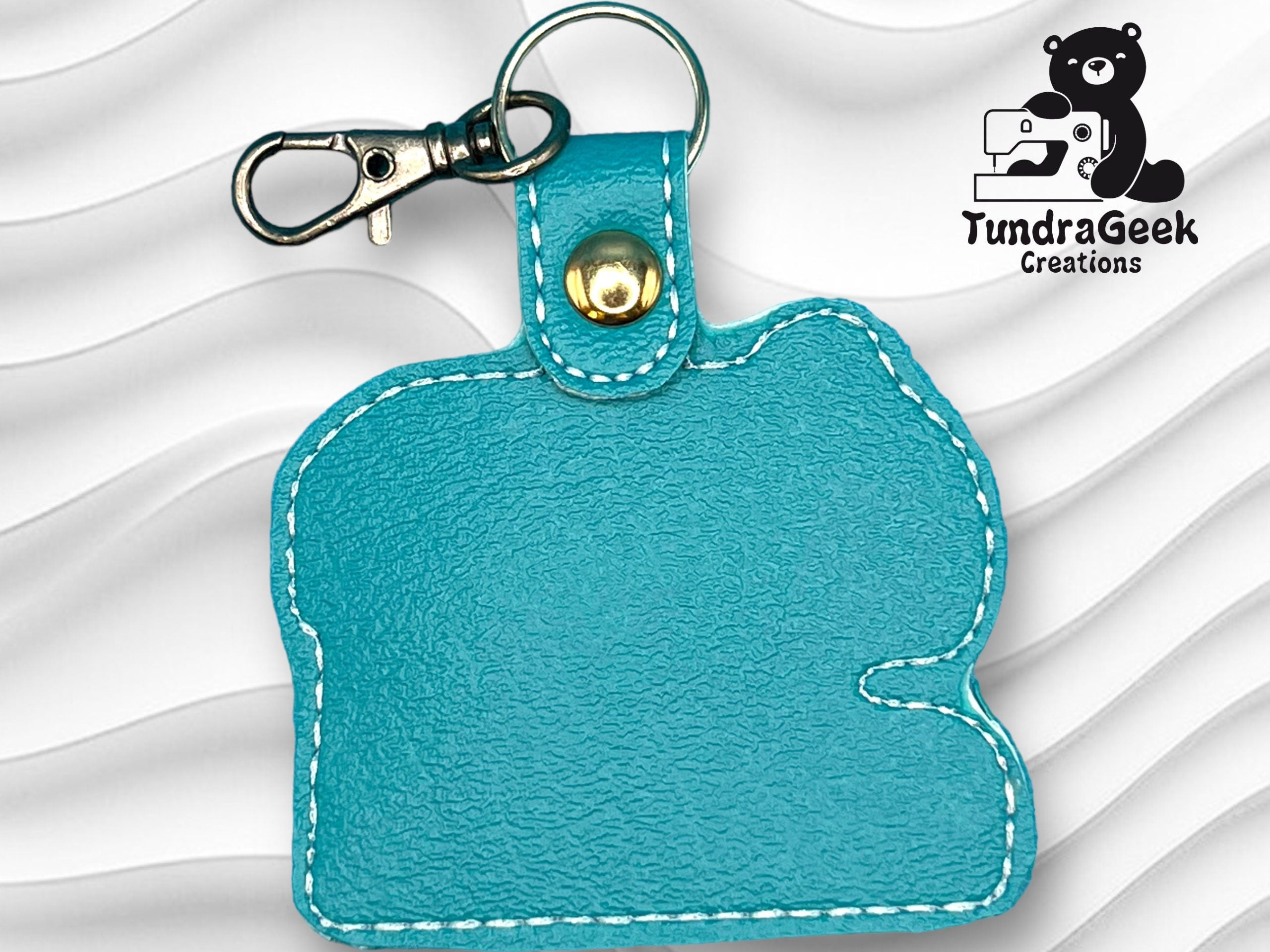 Turquoise leather keychain with white stitching and a metal clasp, displayed on a textured white background. TundraGeek Creations logo is in the top right corner.