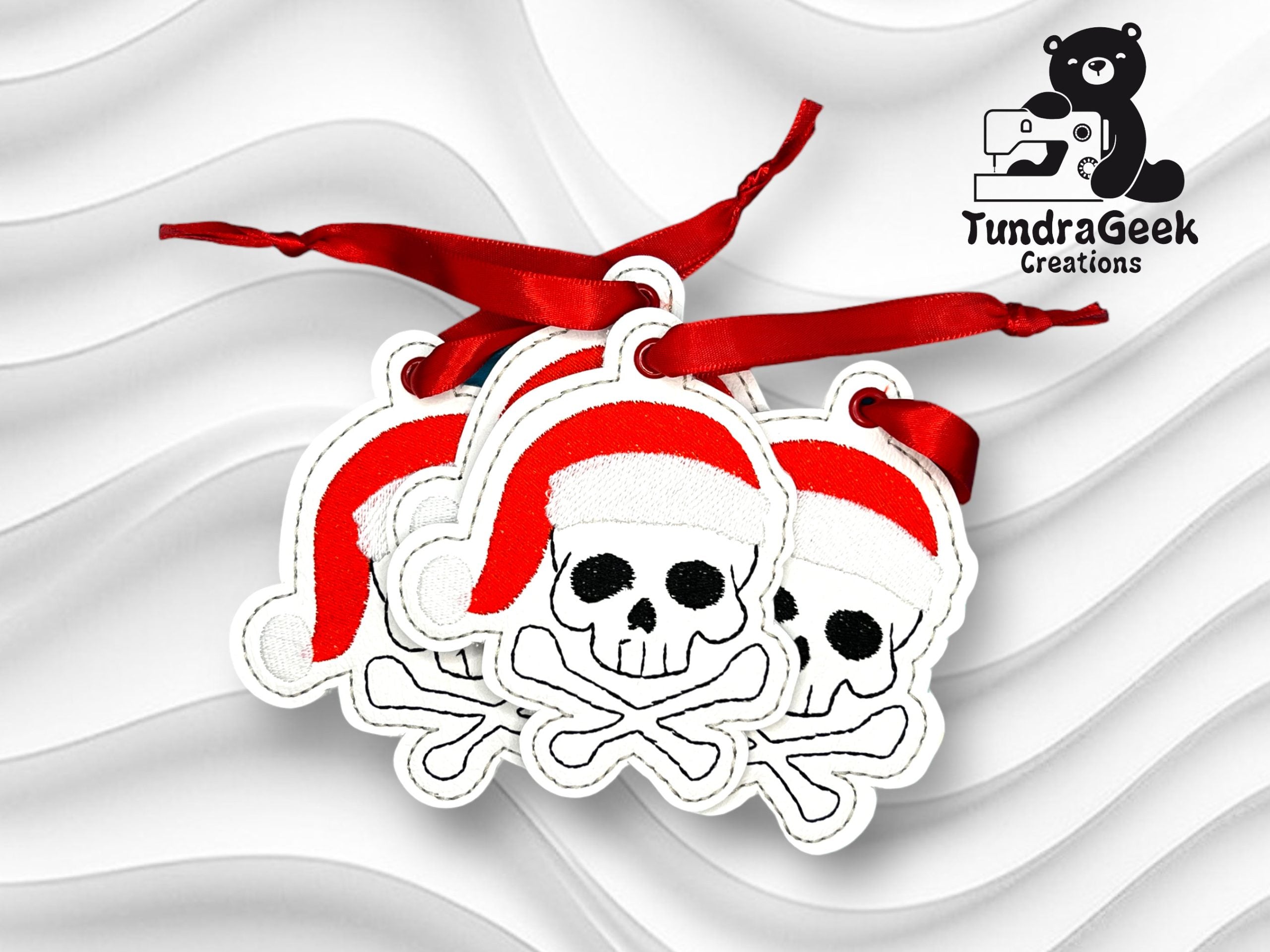 Three skull and crossbones ornaments wearing Santa hats with red ribbons, displayed on a wavy white background with the TundraGeek Creations logo in the top right corner.