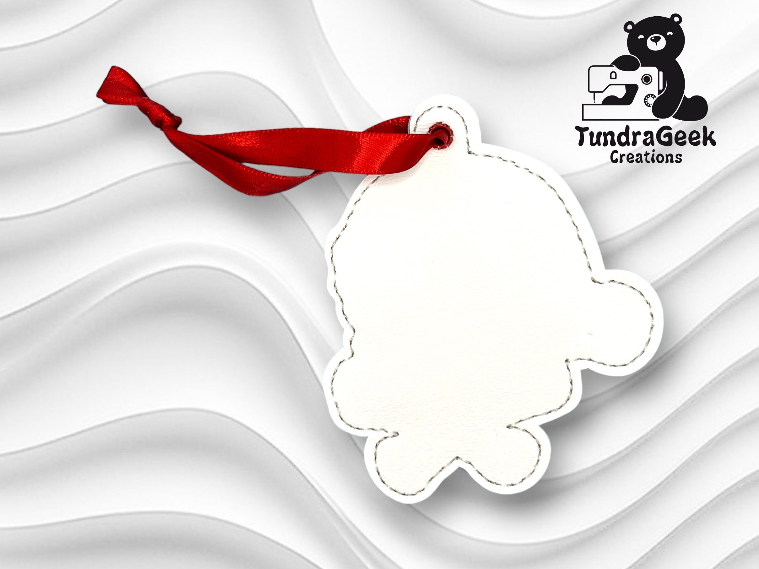A blank, stitched keychain shaped like a character, attached to a red ribbon, with the TundraGeek Creations logo in the top right corner.