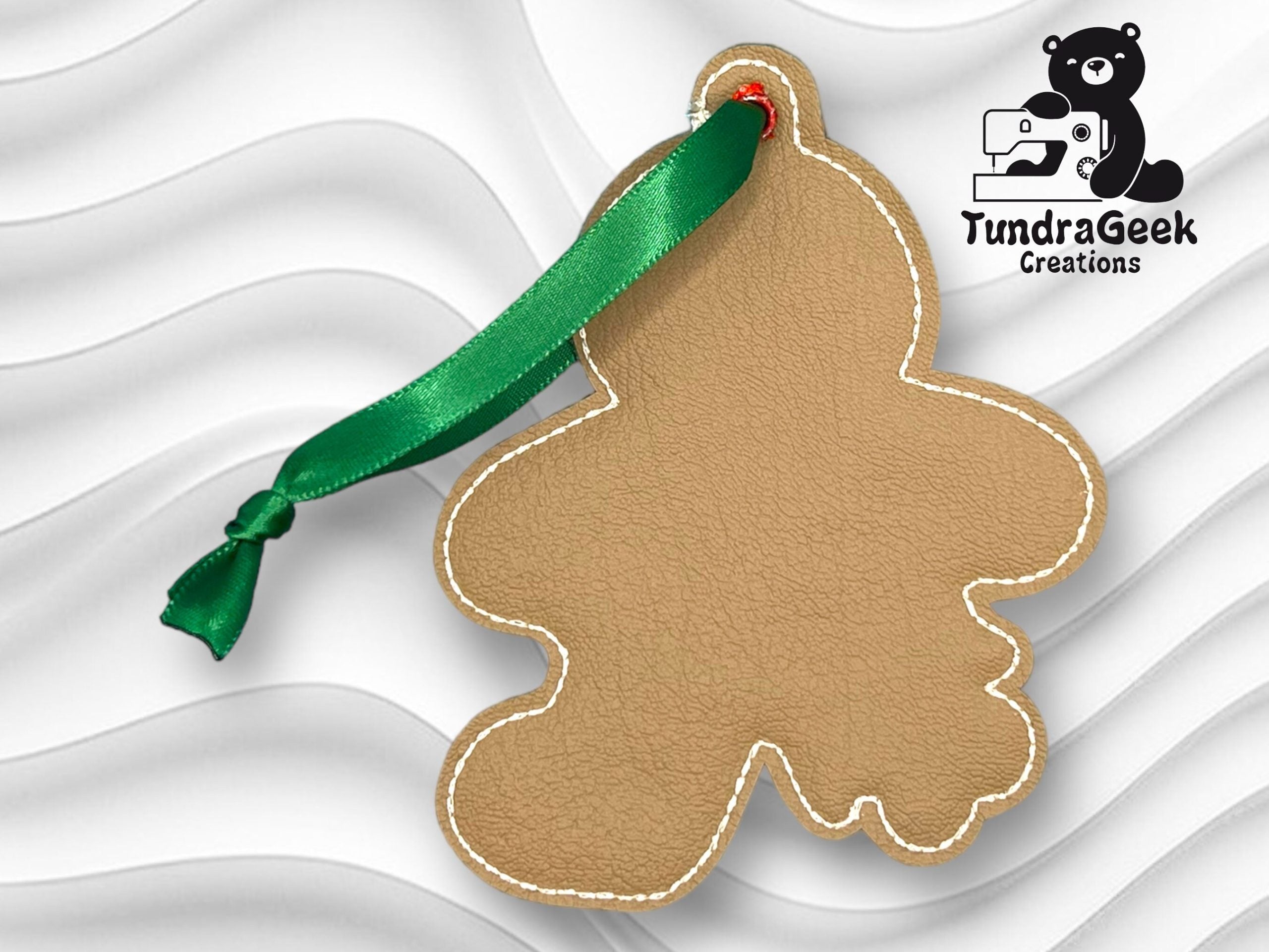 Brown, blank gingerbread person-shaped leather ornament with a green ribbon, shown on a wavy white background with the TundraGeek Creations logo in the corner.