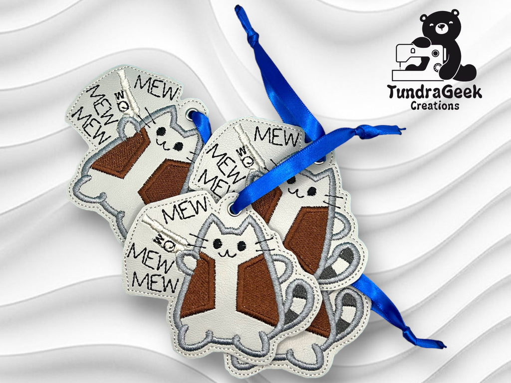 Four cat-shaped keychains with MEW text, made of stitched fabric, are shown with blue ribbons. A logo for TundraGeek Creations featuring a bear and sewing machine is in the top right corner.