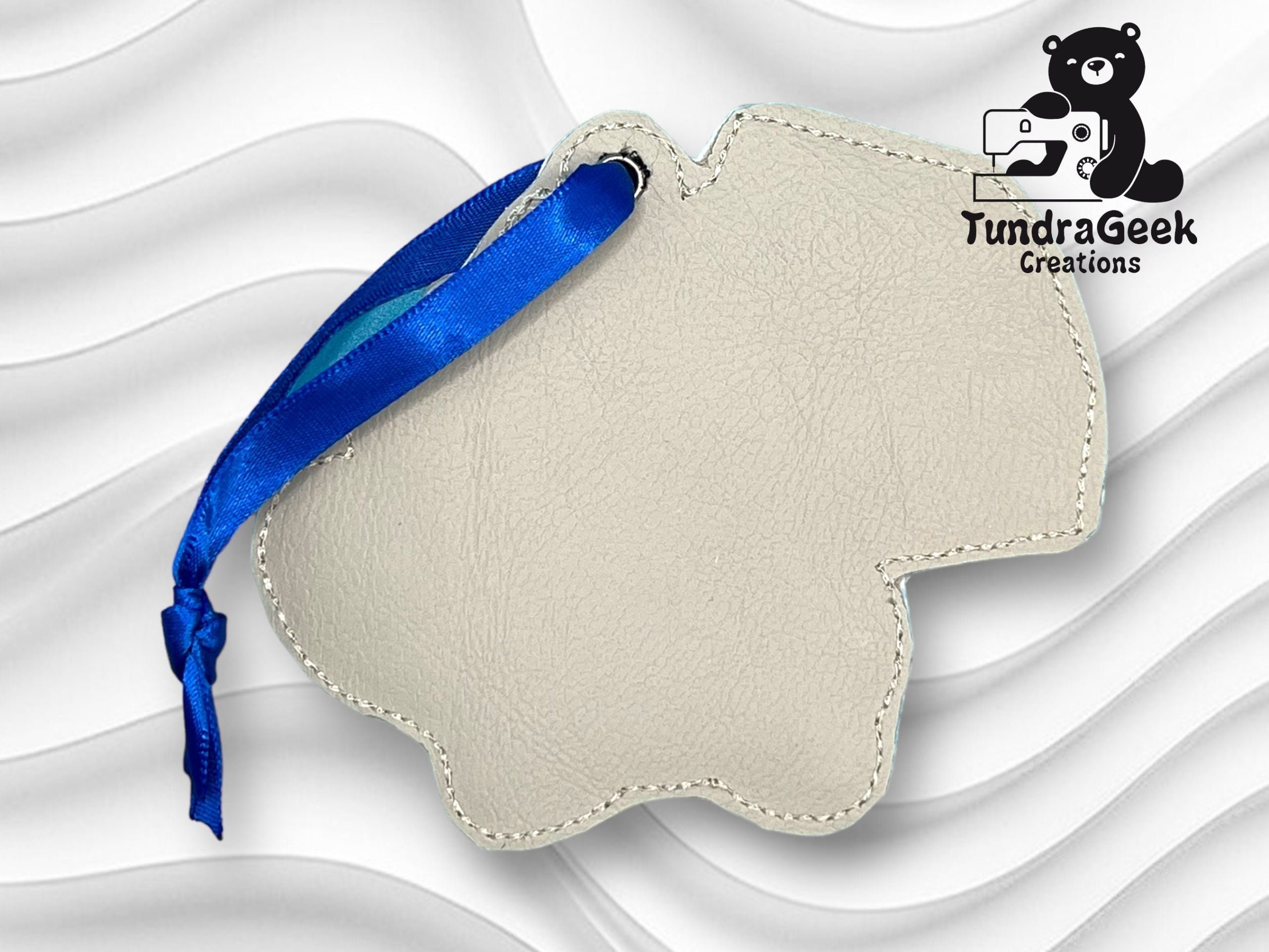 A beige, bear-shaped leather keychain with a blue ribbon, featuring the TundraGeek Creations logo in the top right corner on a wavy white background.