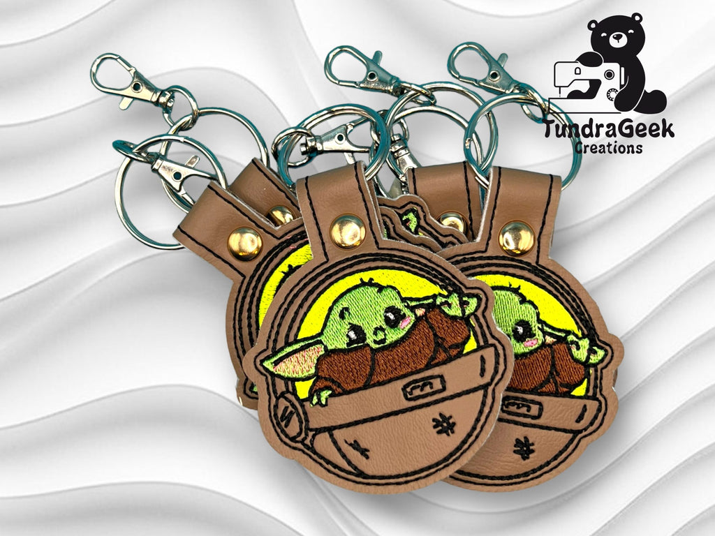 Three brown leather keychains with embroidered green alien characters in pods, displayed overlapping on a wavy white background with the TundraGeek Creations logo in the corner.