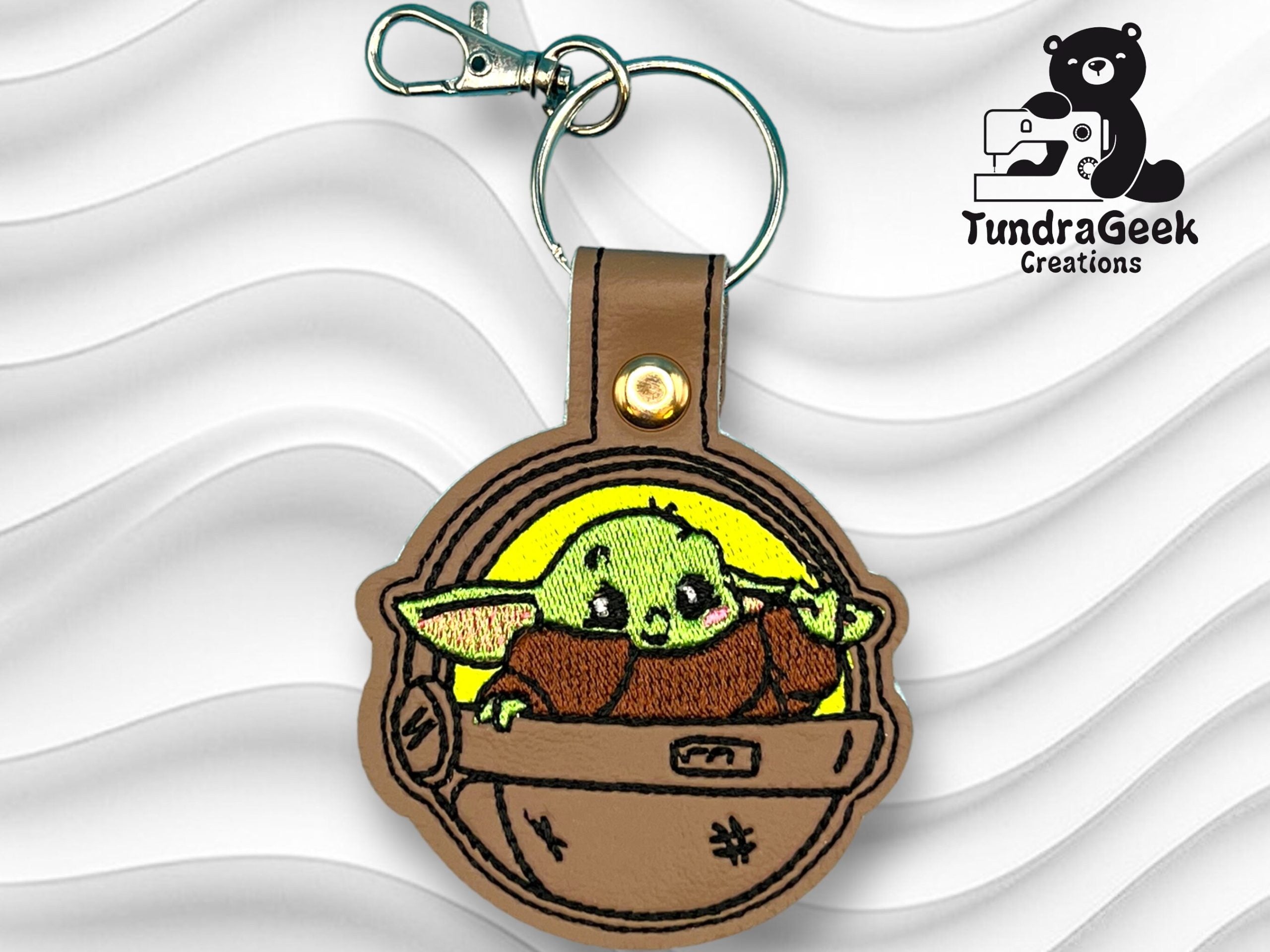 Embroidered keychain featuring a green, cartoonish alien in a brown pod with a metal clasp, displayed on a wavy white background with TundraGeek Creations logo.