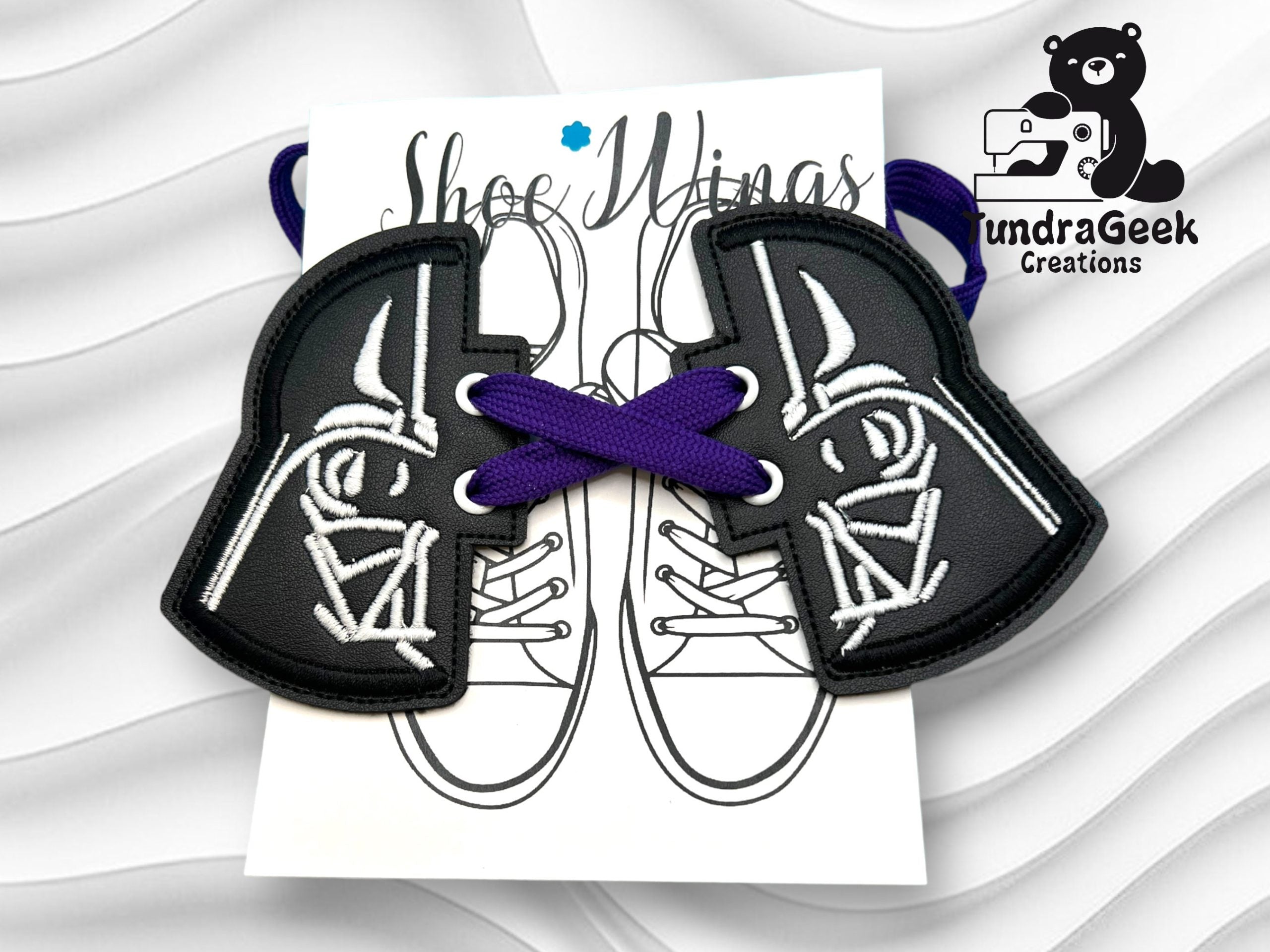 A pair of black Darth Vader-themed shoe accessories with purple laces displayed on a card featuring an outline of sneakers, with the TundraGeek Creations logo in the corner.