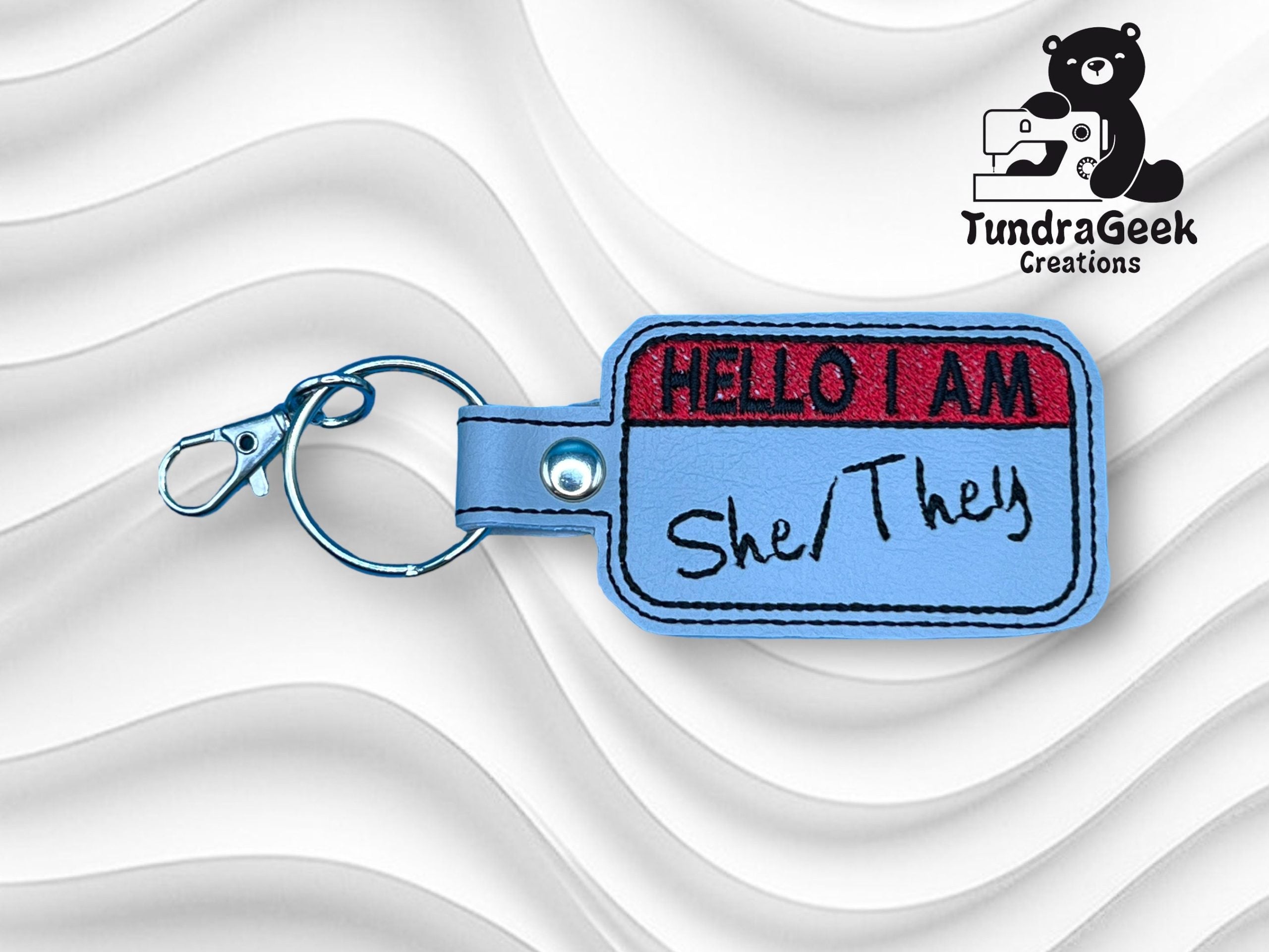 A keychain shaped like a name tag with the text HELLO I AM She/They in handwritten style, displayed on a wavy white background with the TundraGeek Creations logo in the corner.