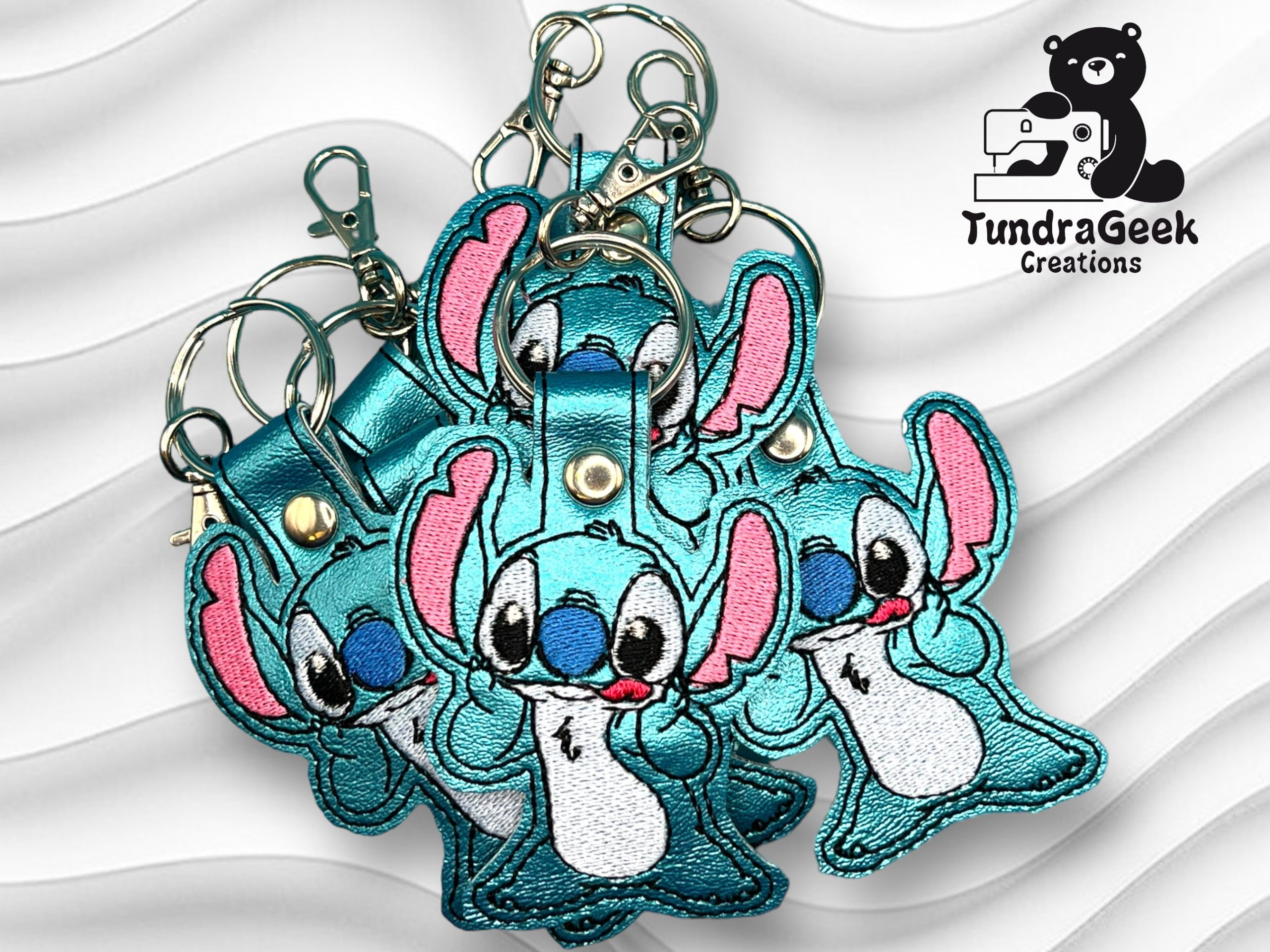 Four embroidered blue cartoon character keychains with pink ears are displayed on a wavy white background, with the TundraGeek Creations logo in the top right corner.