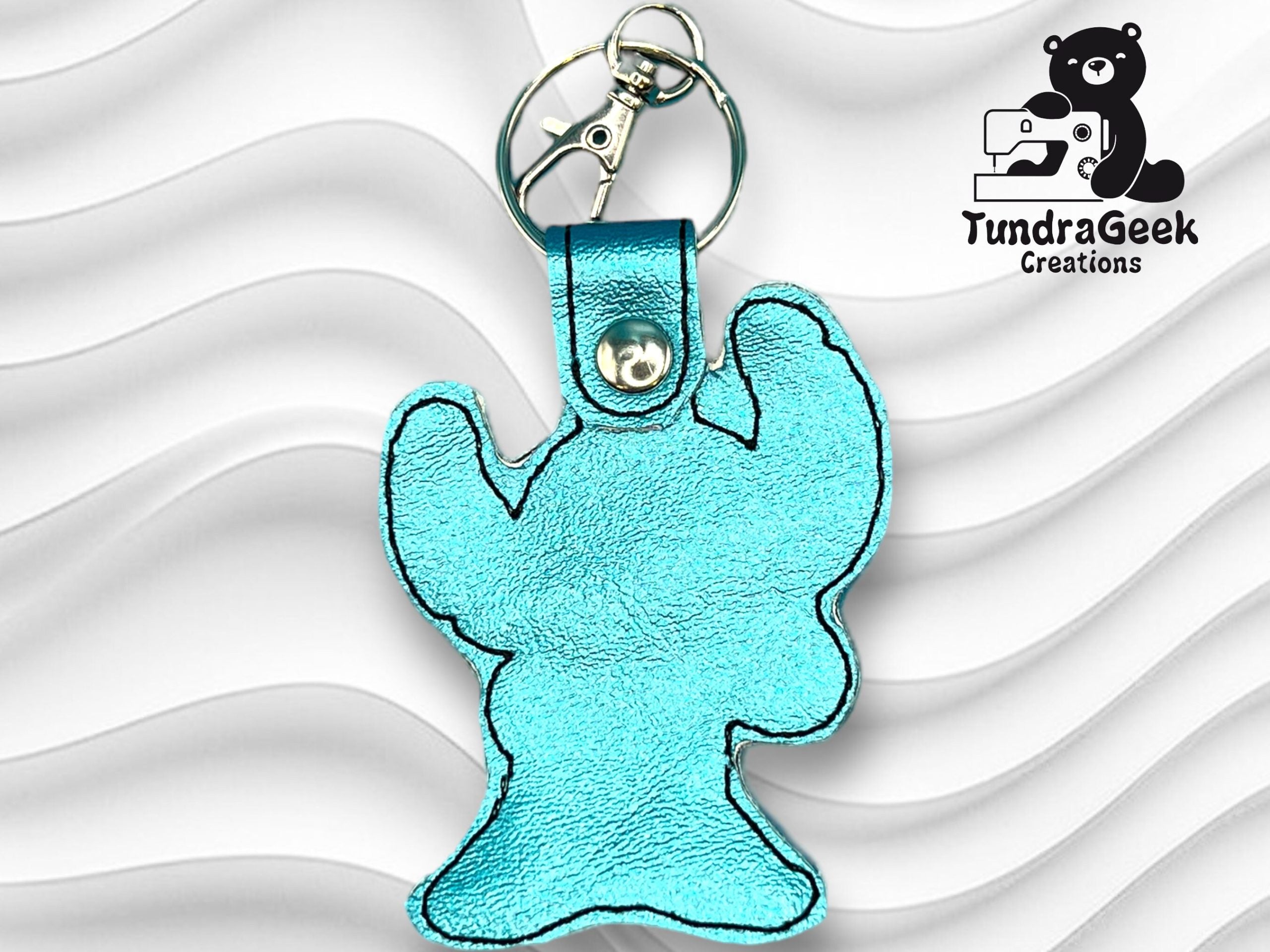 A blue metallic keychain shaped like a cartoon creature is displayed against a white wavy background, with the TundraGeek Creations logo in the top right corner.