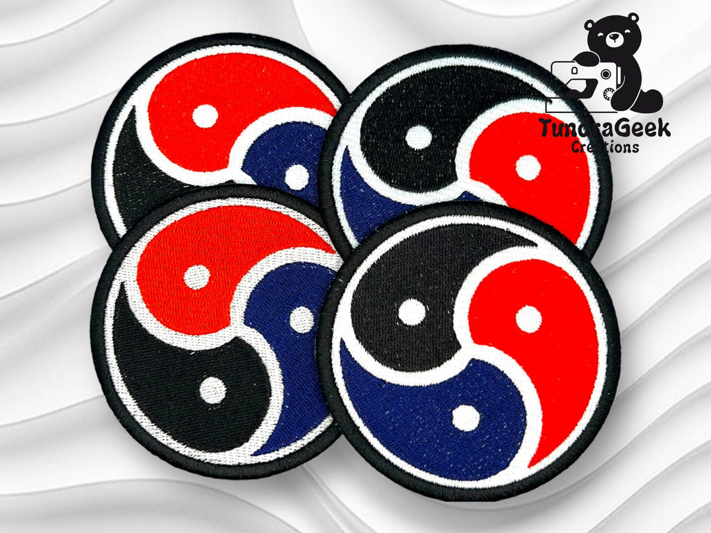 Four round embroidered patches with a tricolor yin-yang design in red, blue, and black are displayed on a wavy white background with a TundraGeek Creations logo in the corner.