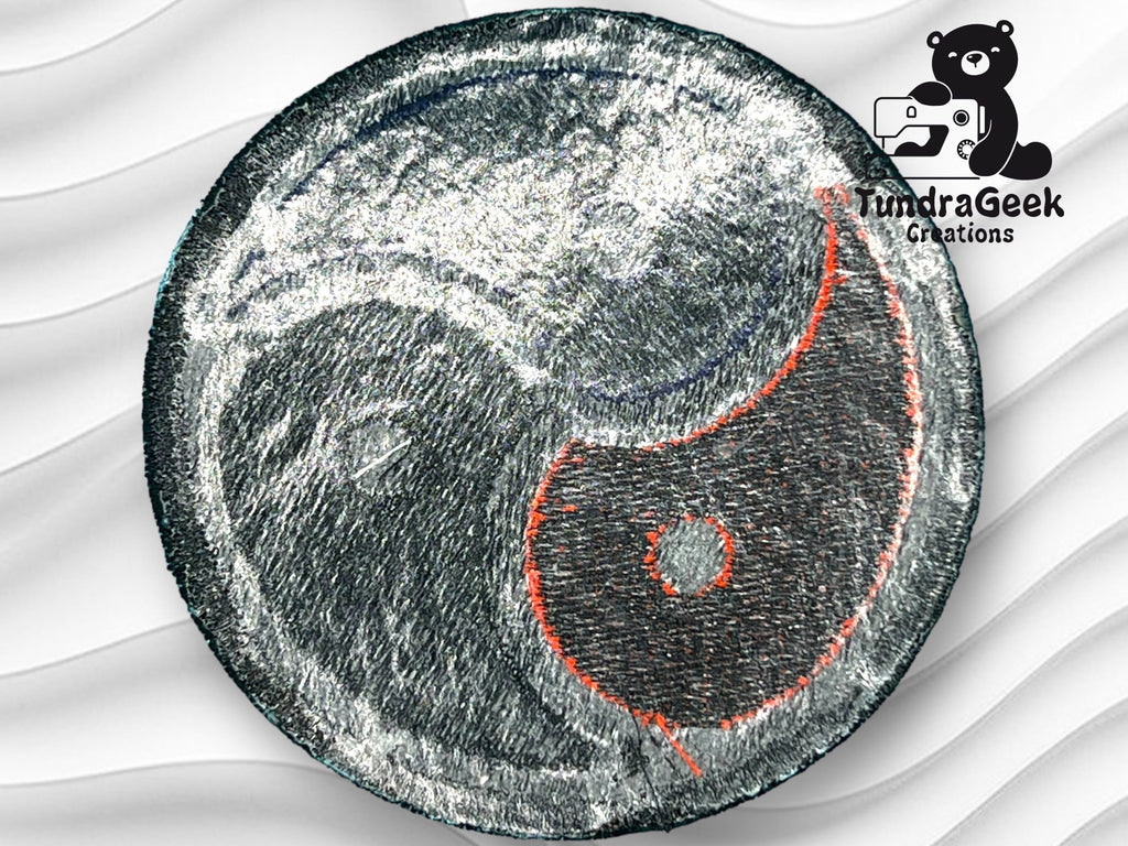 A fabric patch with a yin-yang design featuring metallic silver and black sections, accented with red stitching. The TundraGeek Creations logo appears in the corner.