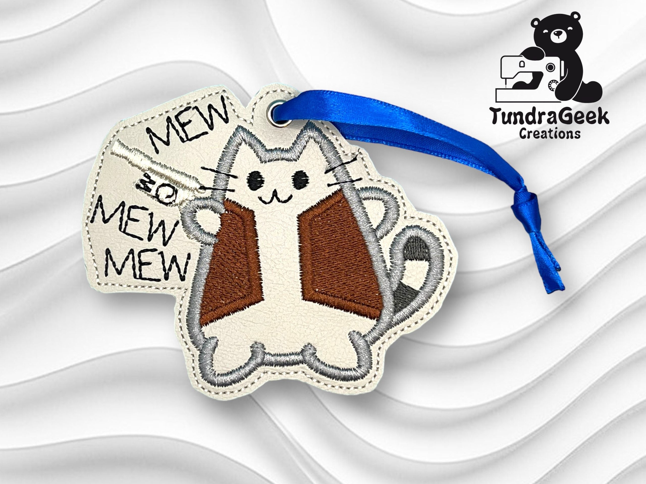 Embroidered keychain featuring a cartoon cat holding a sign that says MEW SO MEW MEW; blue ribbon; TundraGeek Creations logo in the corner.