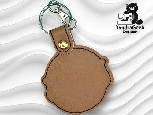 A round brown leather keychain with a snap button and keyring, displayed on a wavy white background. A TundraGeek Creations logo featuring a bear is in the top right corner.