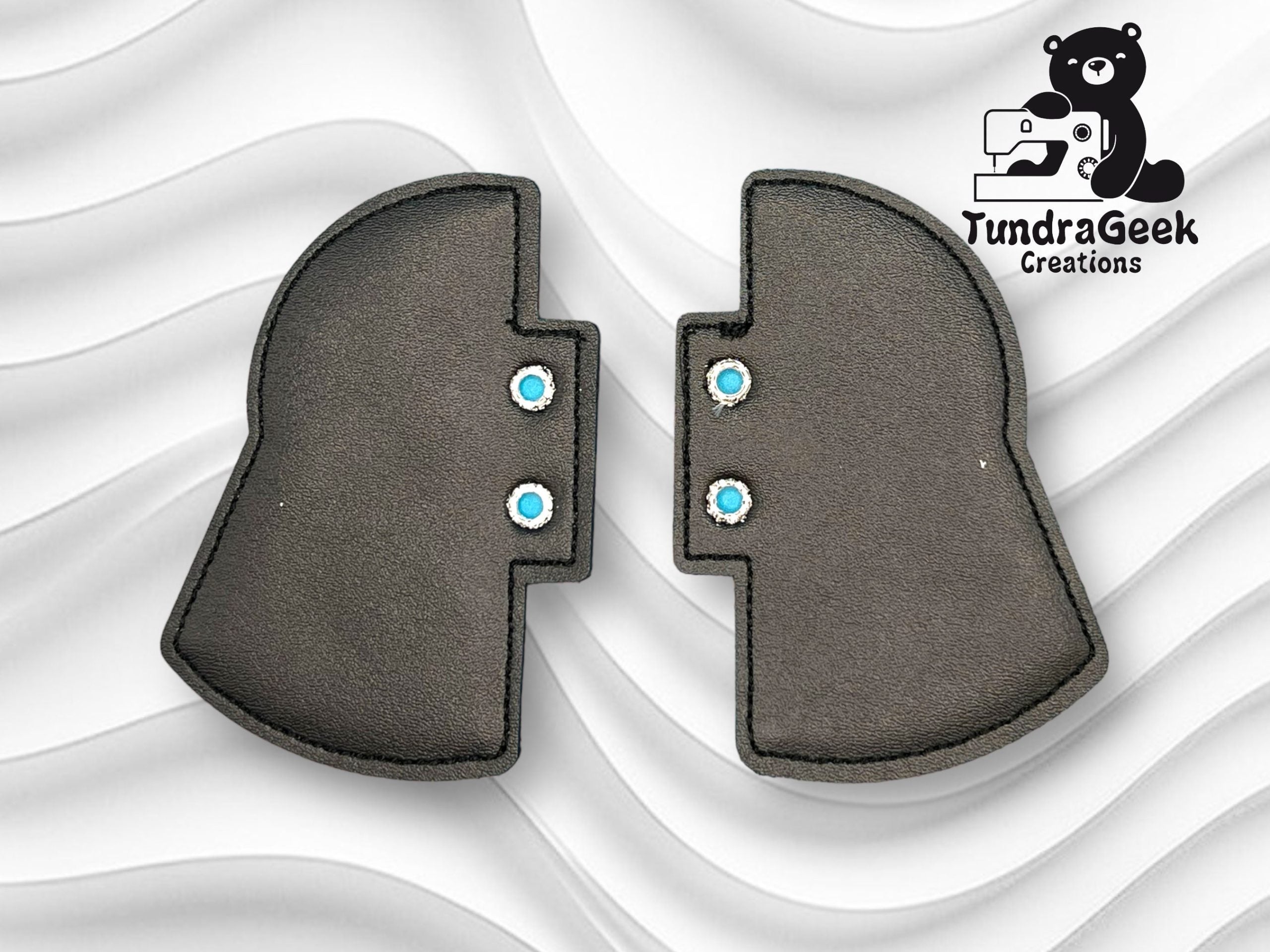 Two black, helmet-shaped pieces with blue snaps are placed side by side on a wavy white background. A TundraGeek Creations logo with a bear is in the top right corner.