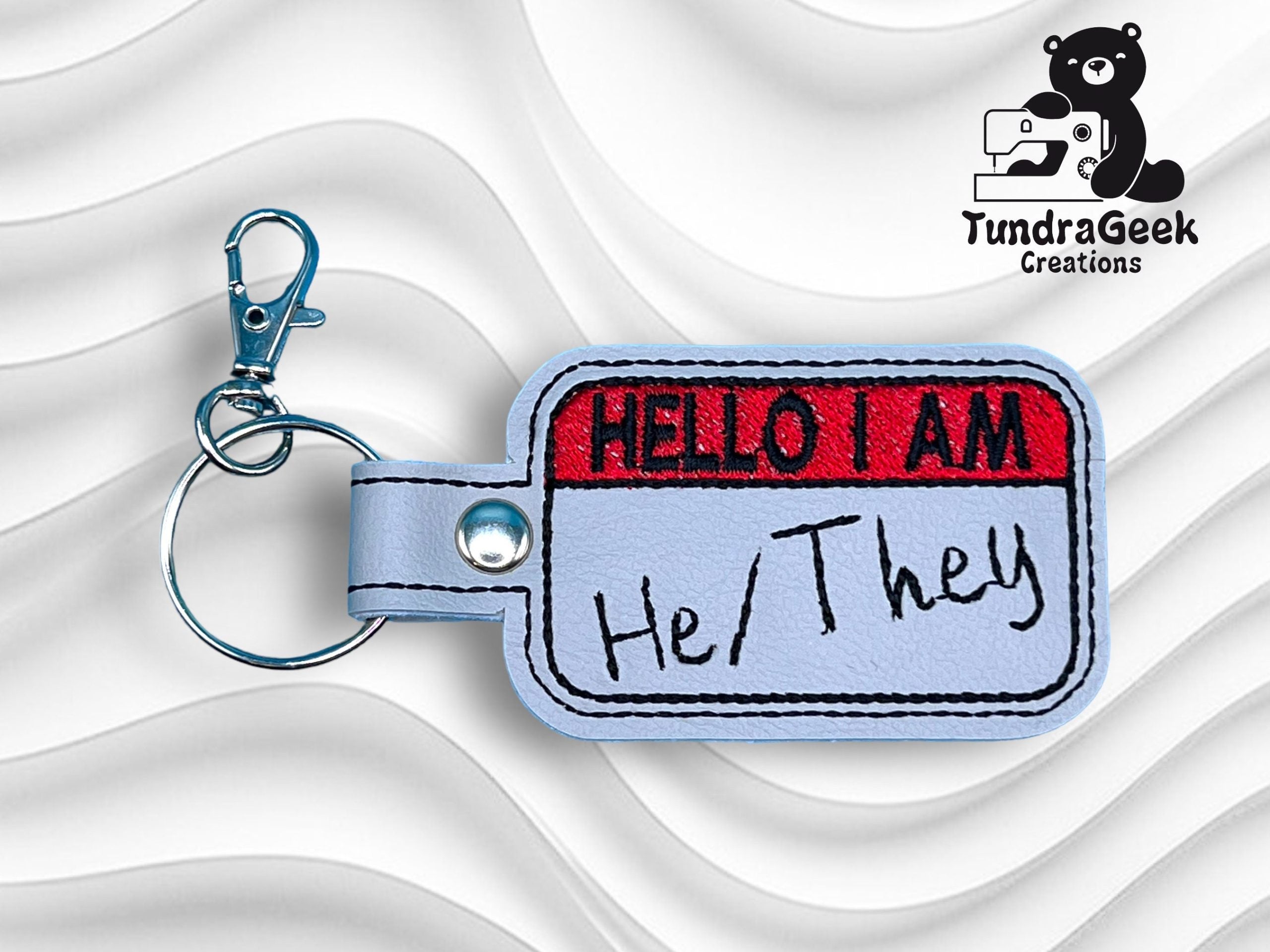 A keychain with a Hello I Am name tag design embroidered in red and black, showing the pronouns He/They written on it. Logo of TundraGeek Creations in the top right corner.