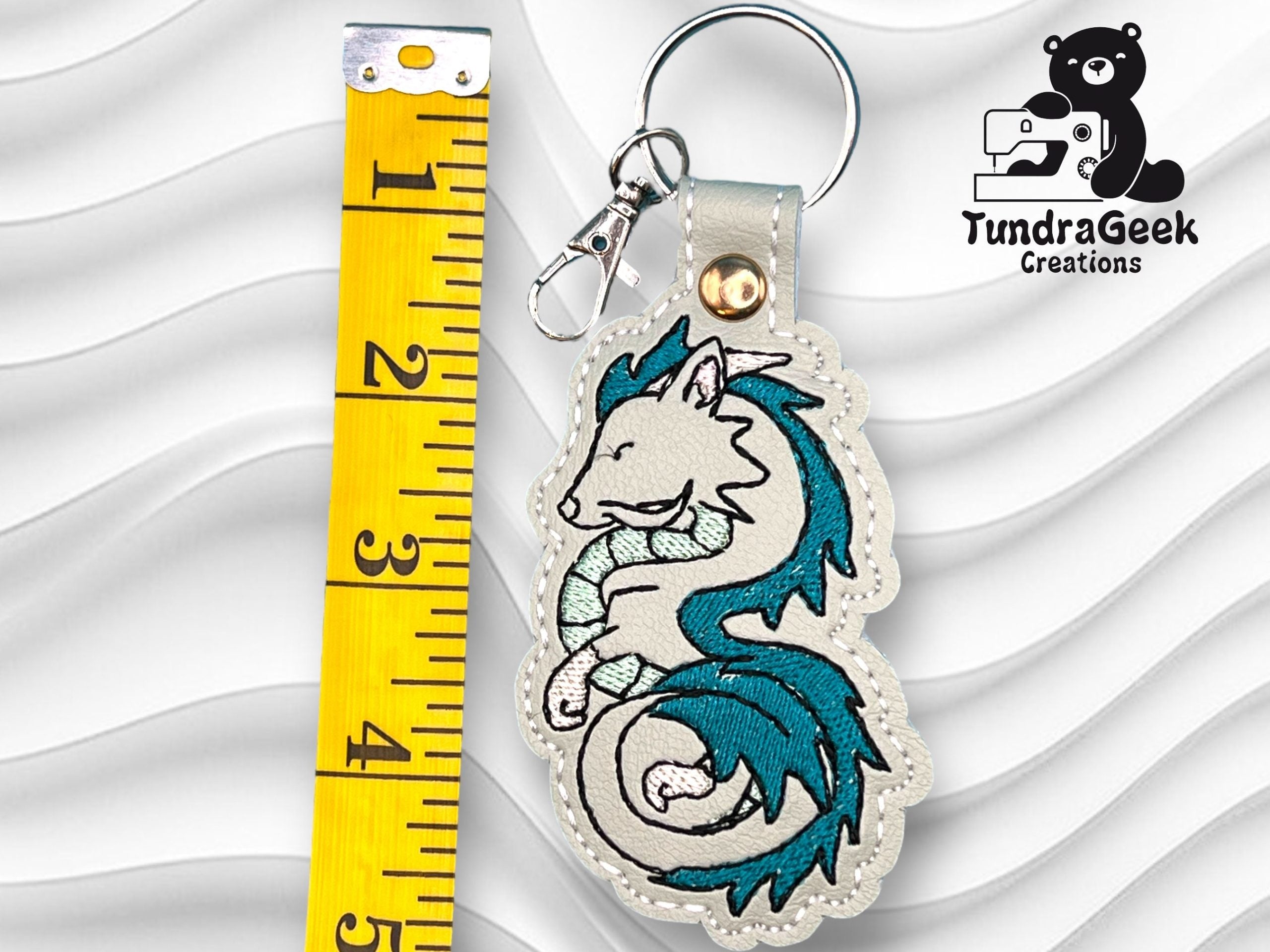Embroidered keychain featuring a curled dragon next to a measuring tape showing it is about 4 inches tall; TundraGeek Creations logo in the corner.