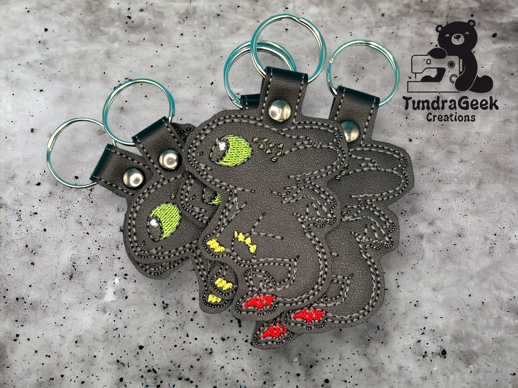 Three black dragon-shaped keychains with embroidered details and colored eyes (green, yellow, red) are displayed on a gray textured surface. Logo TundraGeek Creations is visible.