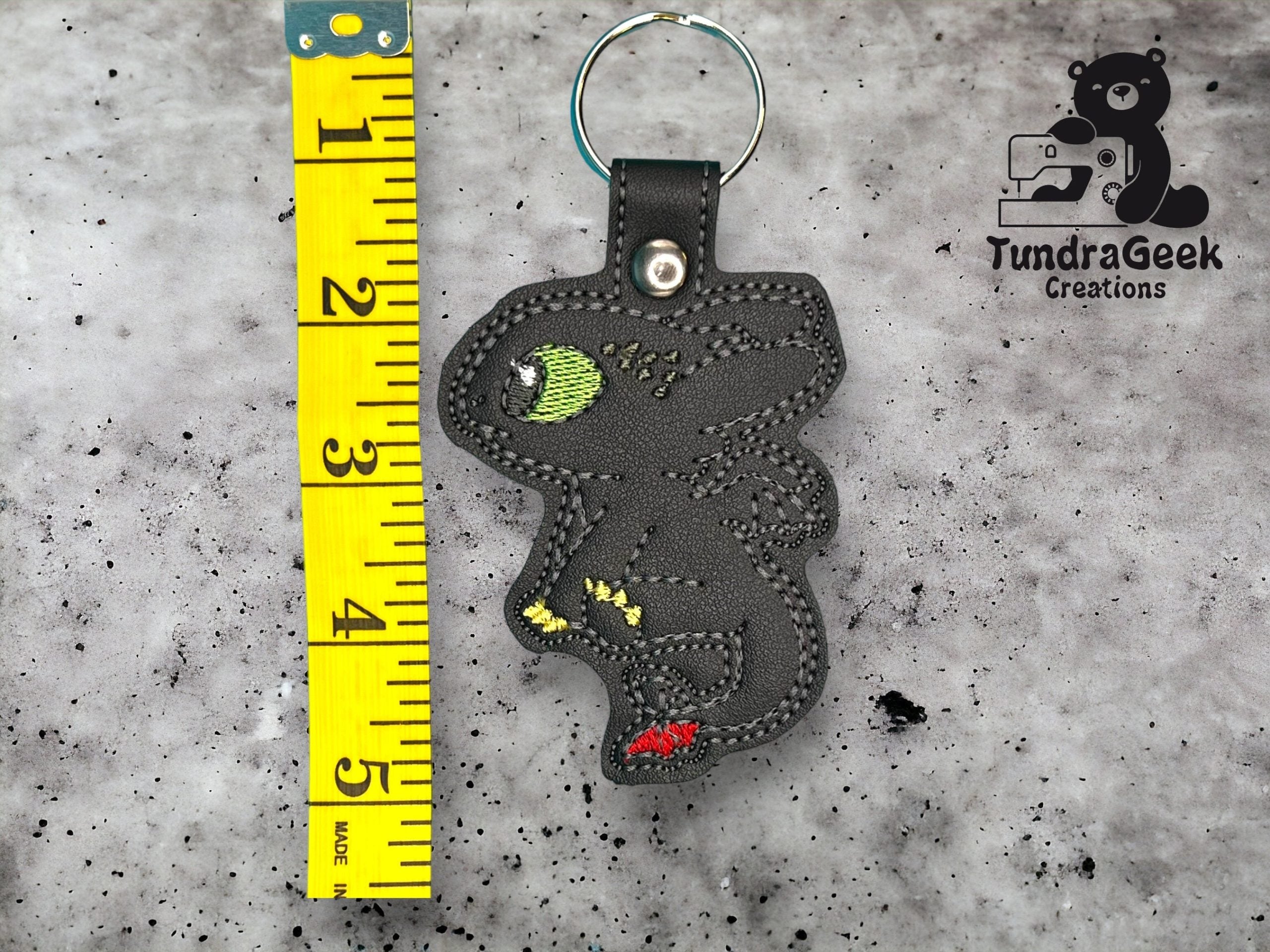 A black dragon keychain with embroidered details is placed next to a measuring tape showing about 4 inches. TundraGeek Creations logo is in the top right corner.