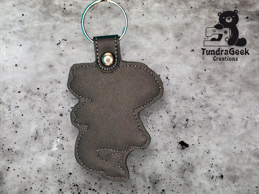 A gray faux leather keychain in the shape of an animal, attached to a metal ring, is displayed on a speckled surface. The TundraGeek Creations logo appears in the top right corner.