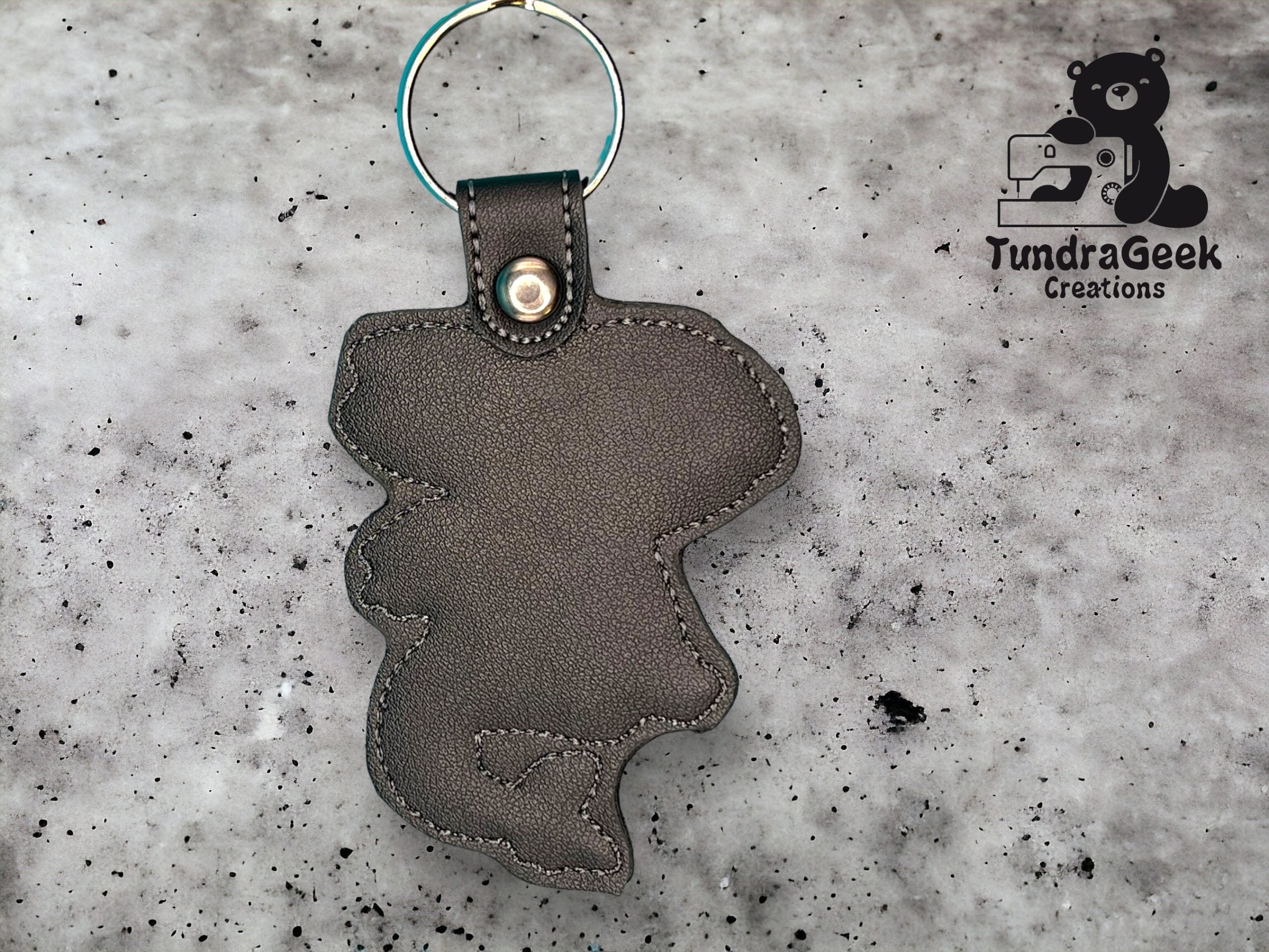 A gray faux leather keychain in the shape of an animal, attached to a metal ring, is displayed on a speckled surface. The TundraGeek Creations logo appears in the top right corner.