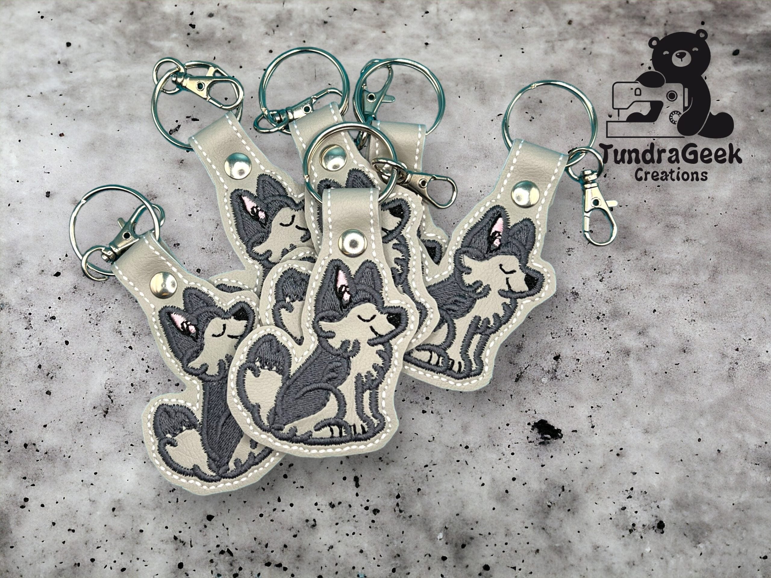 Five keychains with cartoon wolf designs are arranged on a gray speckled background. A TundraGeek Creations logo appears in the top right corner.