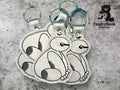 Three silver keychains with stitched cartoon-style animal designs are displayed on a gray surface next to the TundraGeek Creations logo.