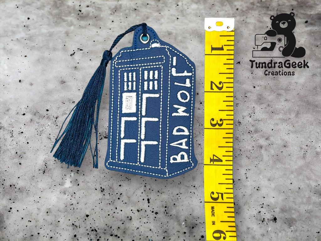 Blue embroidered TARDIS keychain with BAD WOLF text, next to a yellow measuring tape showing a length of about 5 inches. TundraGeek Creations logo is in the top right corner.