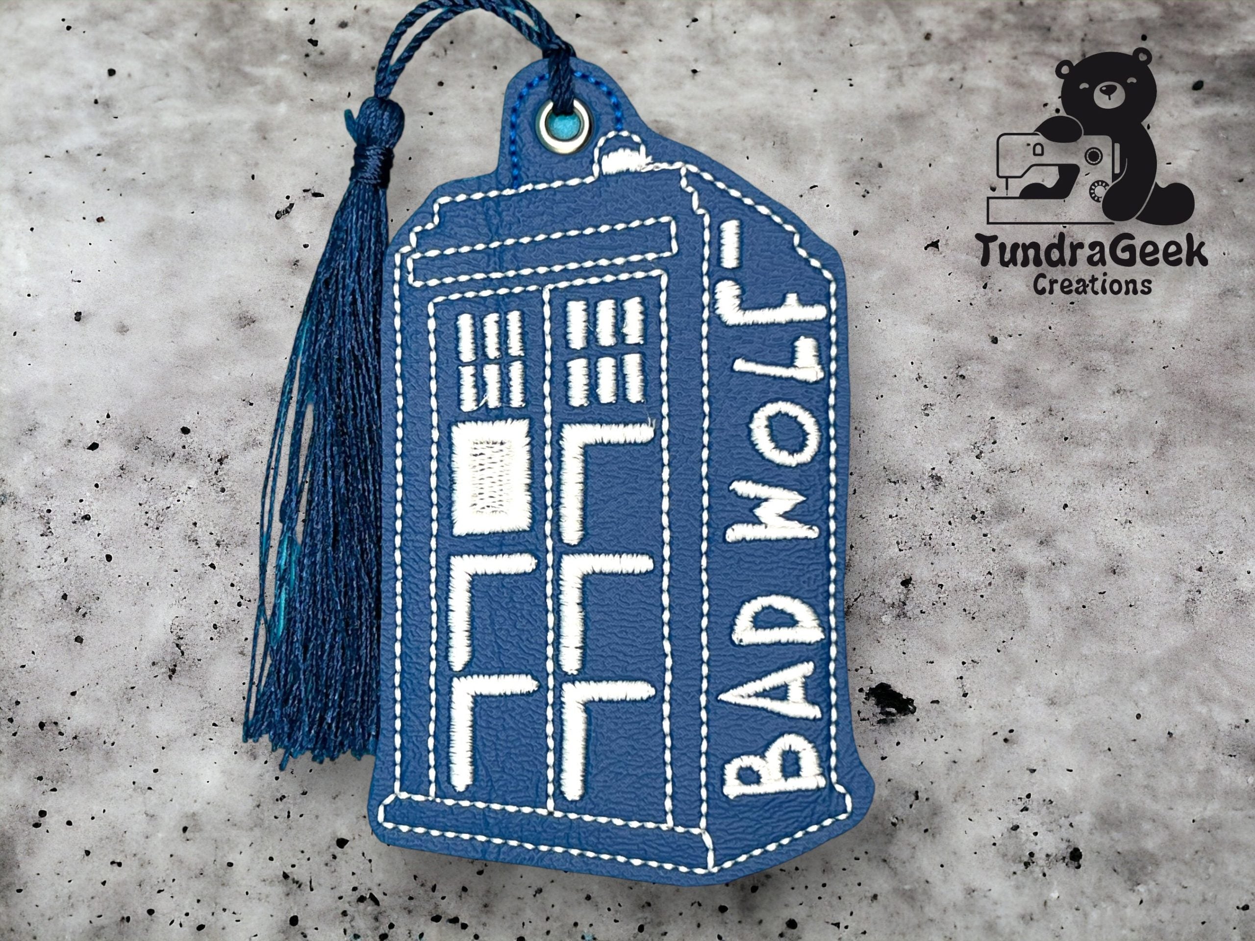A blue, rectangular keychain shaped like the TARDIS from Doctor Who, embroidered with the words BAD WOLF and a blue tassel attached. TundraGeek Creations logo in the corner.