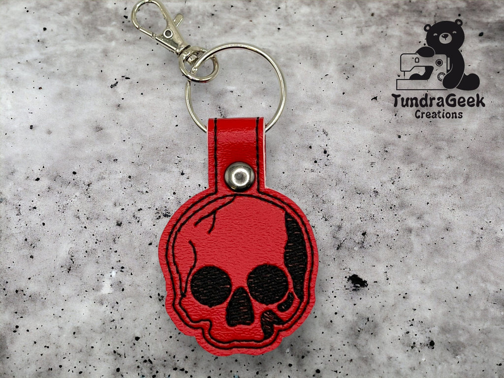 A red skull-shaped leather keychain with a metal ring and clasp is displayed on a gray speckled surface. The TundraGeek Creations logo appears in the upper right corner.
