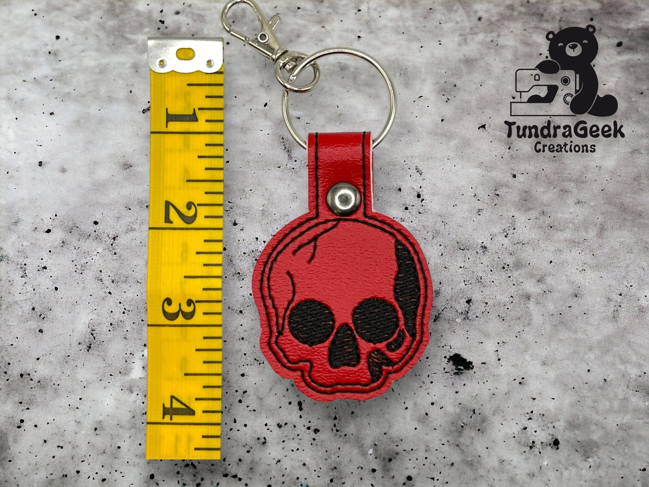 A red skull-shaped keychain with a metal ring and clasp is placed next to a measuring tape showing about 3 inches. The TundraGeek Creations logo is visible in the top right corner.