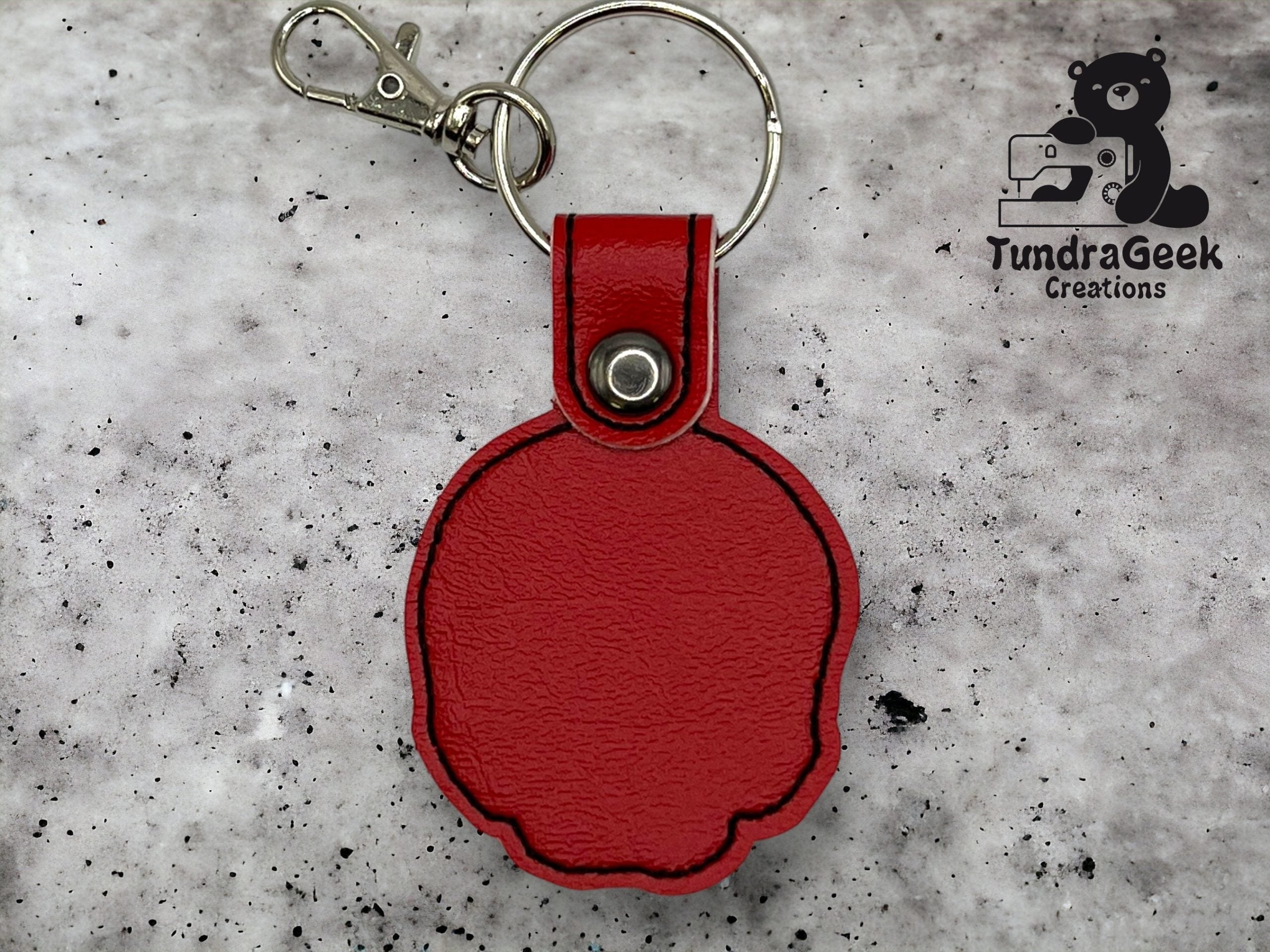 A red faux leather keychain with a snap button and metal clasp is displayed on a speckled background. The TundraGeek Creations logo appears in the top right corner.