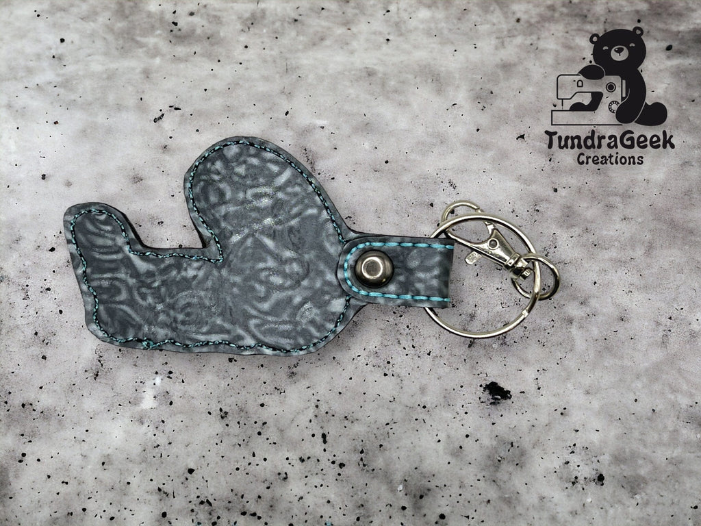 A fabric keychain in the shape of a seal with blue stitching lies on a speckled surface, next to a metal key ring and clip. TundraGeek Creations logo is in the top right corner.