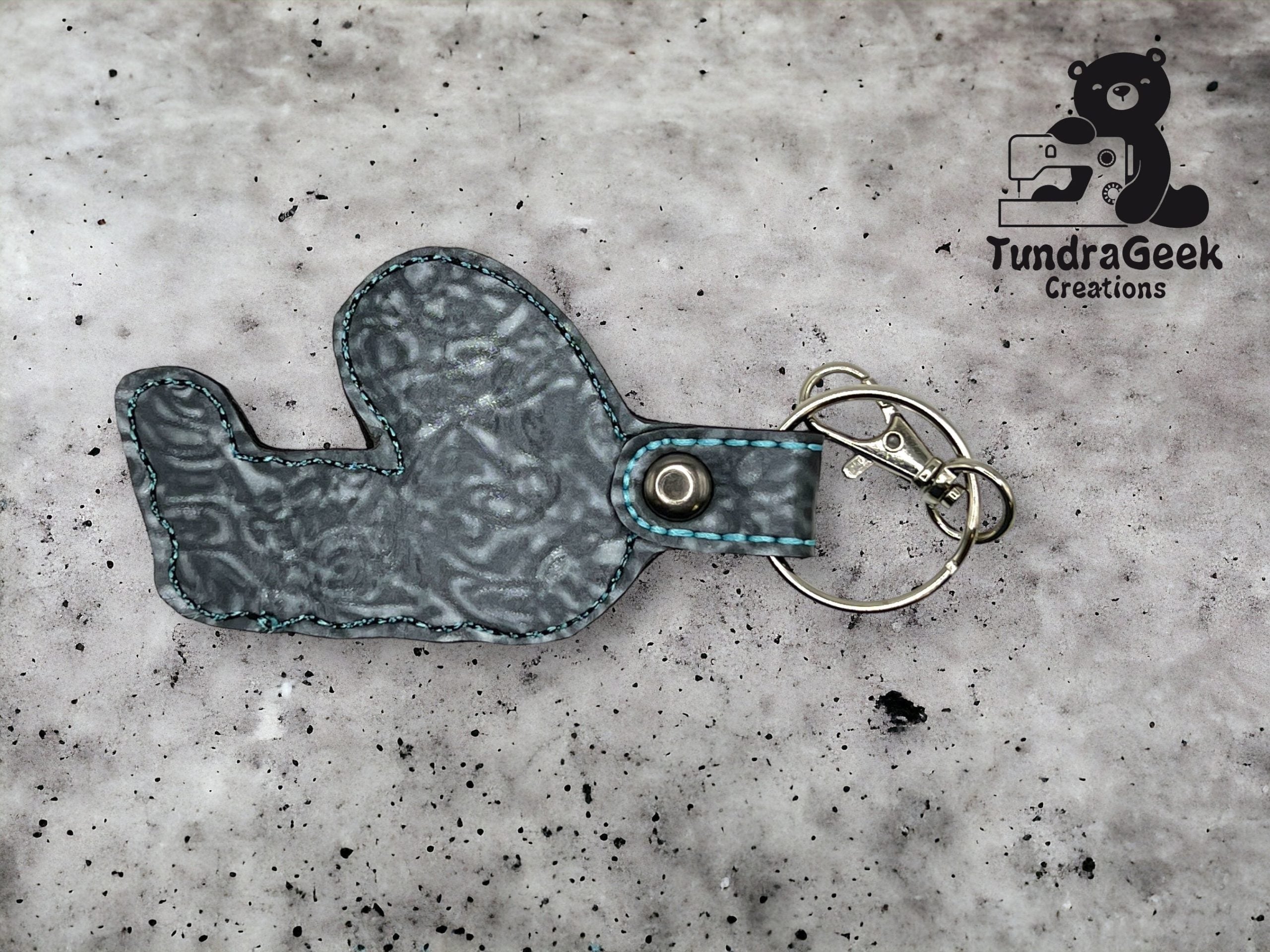 A fabric keychain in the shape of a seal with blue stitching lies on a speckled surface, next to a metal key ring and clip. TundraGeek Creations logo is in the top right corner.