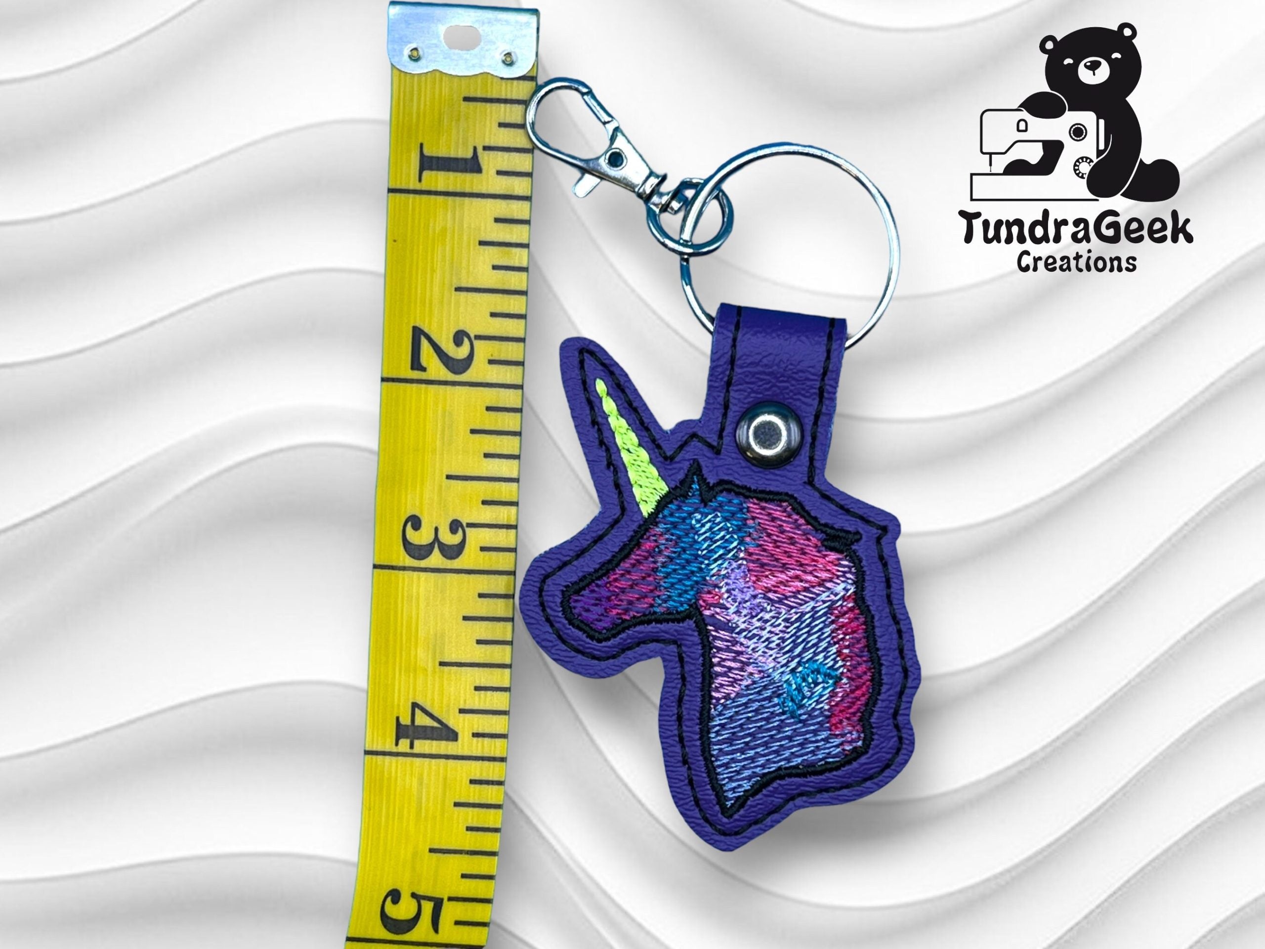 A keychain with an embroidered unicorn head next to a yellow measuring tape showing about 3.5 inches; TundraGeek Creations logo is in the top right corner.