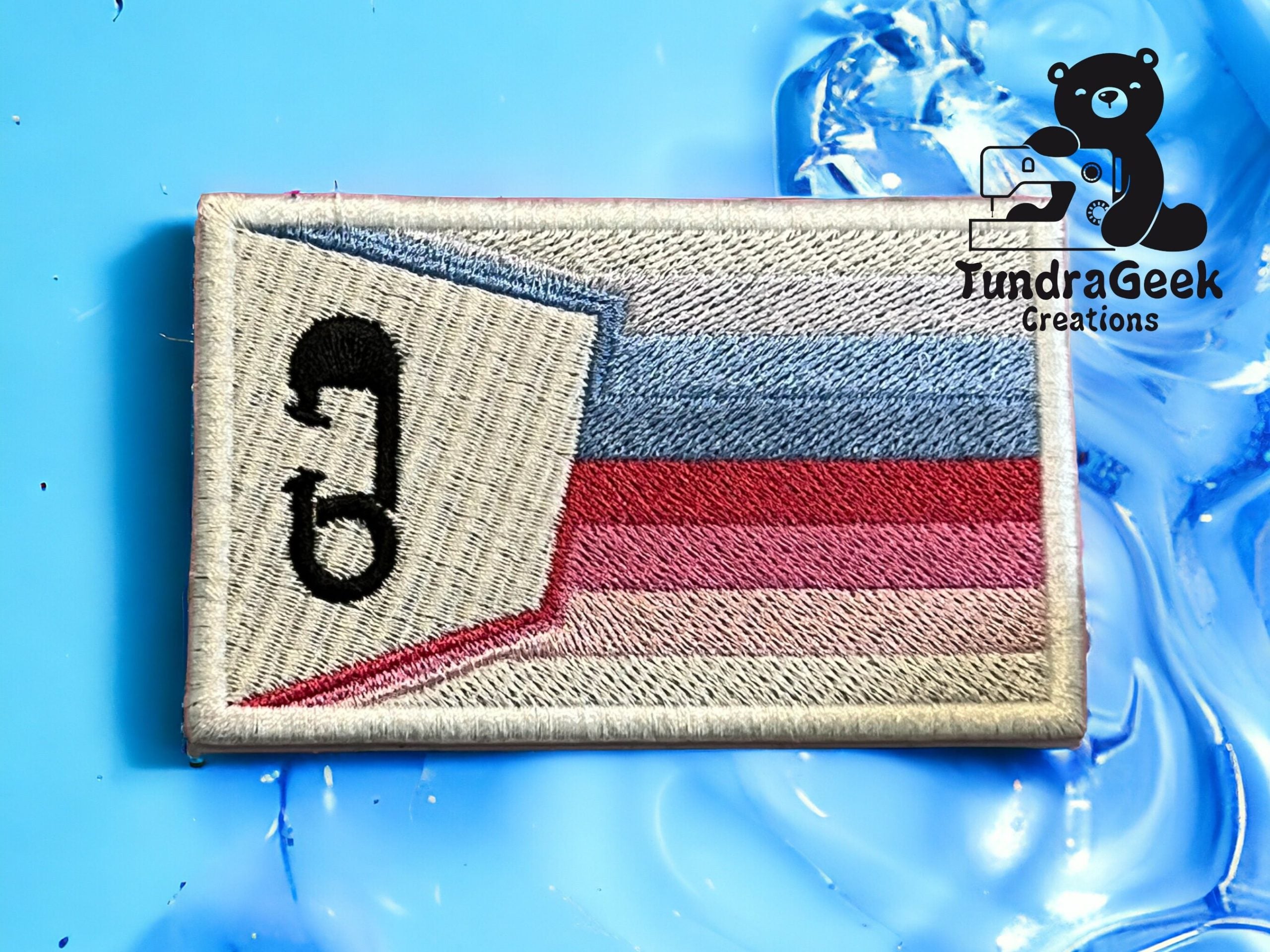 Rectangular embroidered patch with horizontal blue, white, and pink stripes, a black stylized safety pin, and the TundraGeek Creations logo on a blue ice-like background.