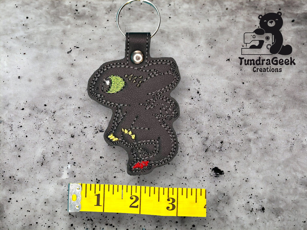 A black dragon-shaped keychain with green and red details lies next to a measuring tape on a gray surface. The logo TundraGeek Creations appears in the top right corner.