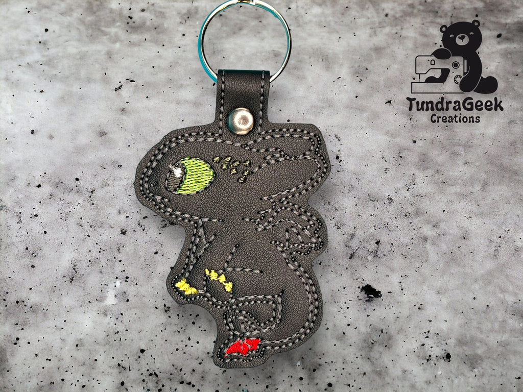 A black faux leather keychain shaped like a stylized dragon with green, yellow, and red details, placed on a speckled gray background. TundraGeek Creations logo is visible.