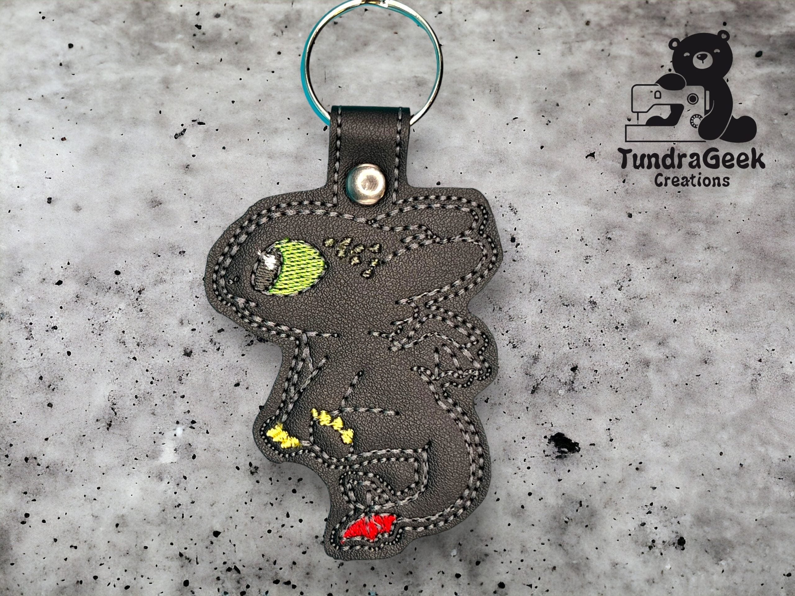 A black faux leather keychain shaped like a stylized dragon with green, yellow, and red details, placed on a speckled gray background. TundraGeek Creations logo is visible.