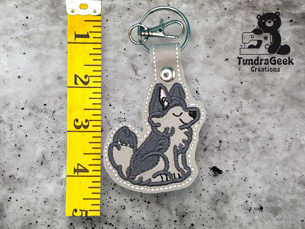 A keychain shaped like a gray wolf with a pink bow is placed next to a yellow measuring tape showing a length of about 4 inches. The TundraGeek Creations logo is in the corner.