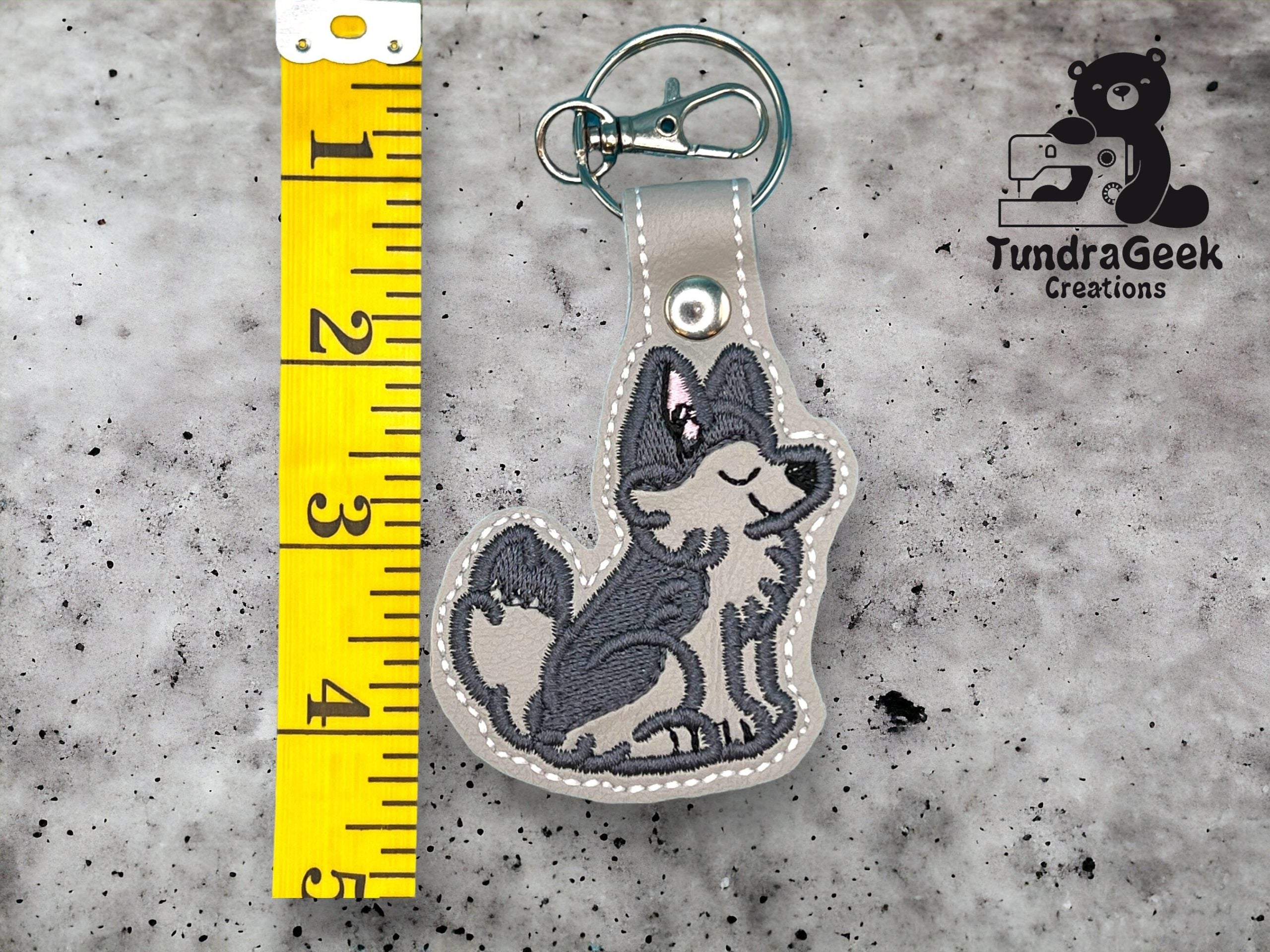 A keychain shaped like a gray wolf with a pink bow is placed next to a yellow measuring tape showing a length of about 4 inches. The TundraGeek Creations logo is in the corner.