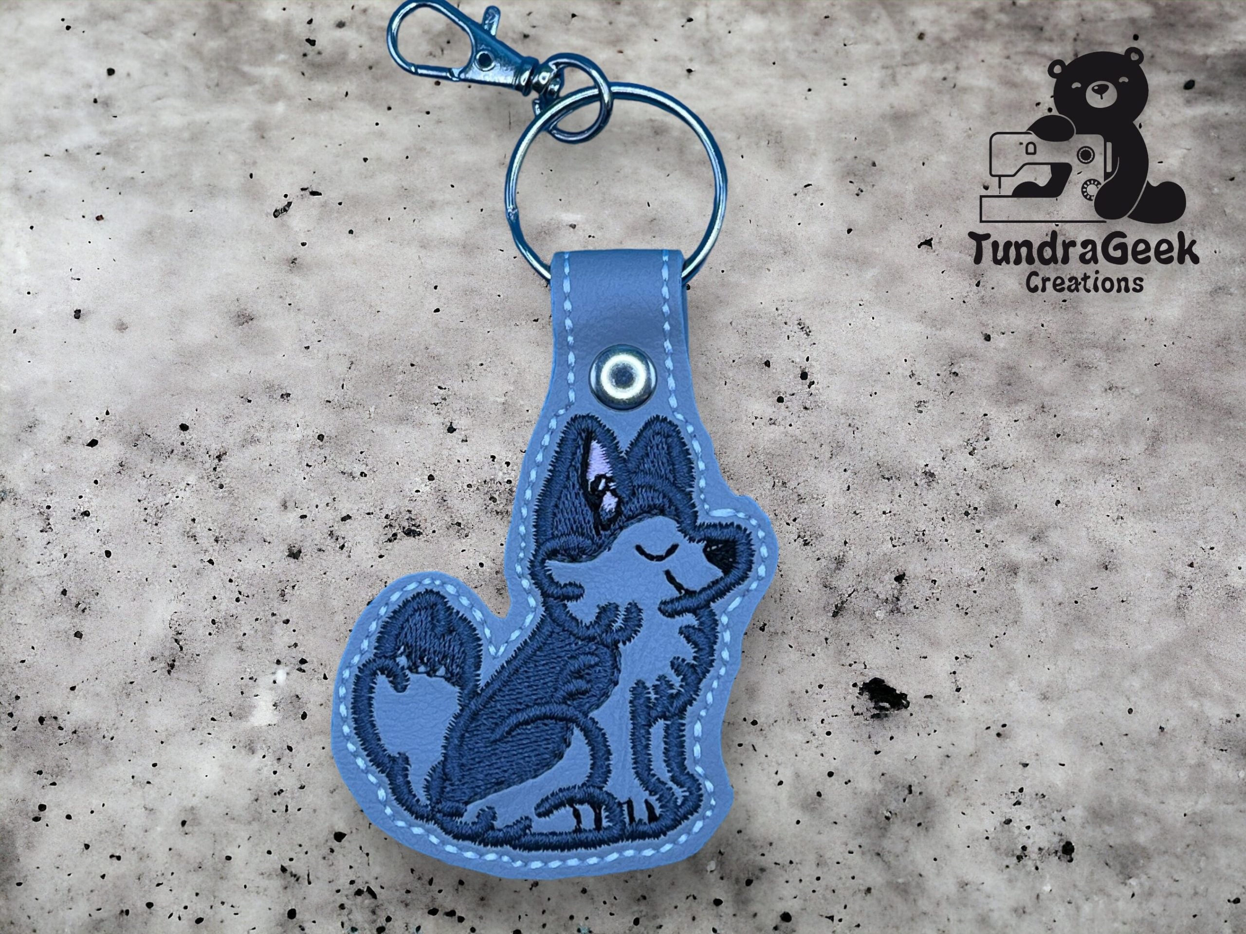 Blue wolf-shaped keychain with embroidered details and a metal clip, displayed on a textured surface with the TundraGeek Creations logo in the background.
