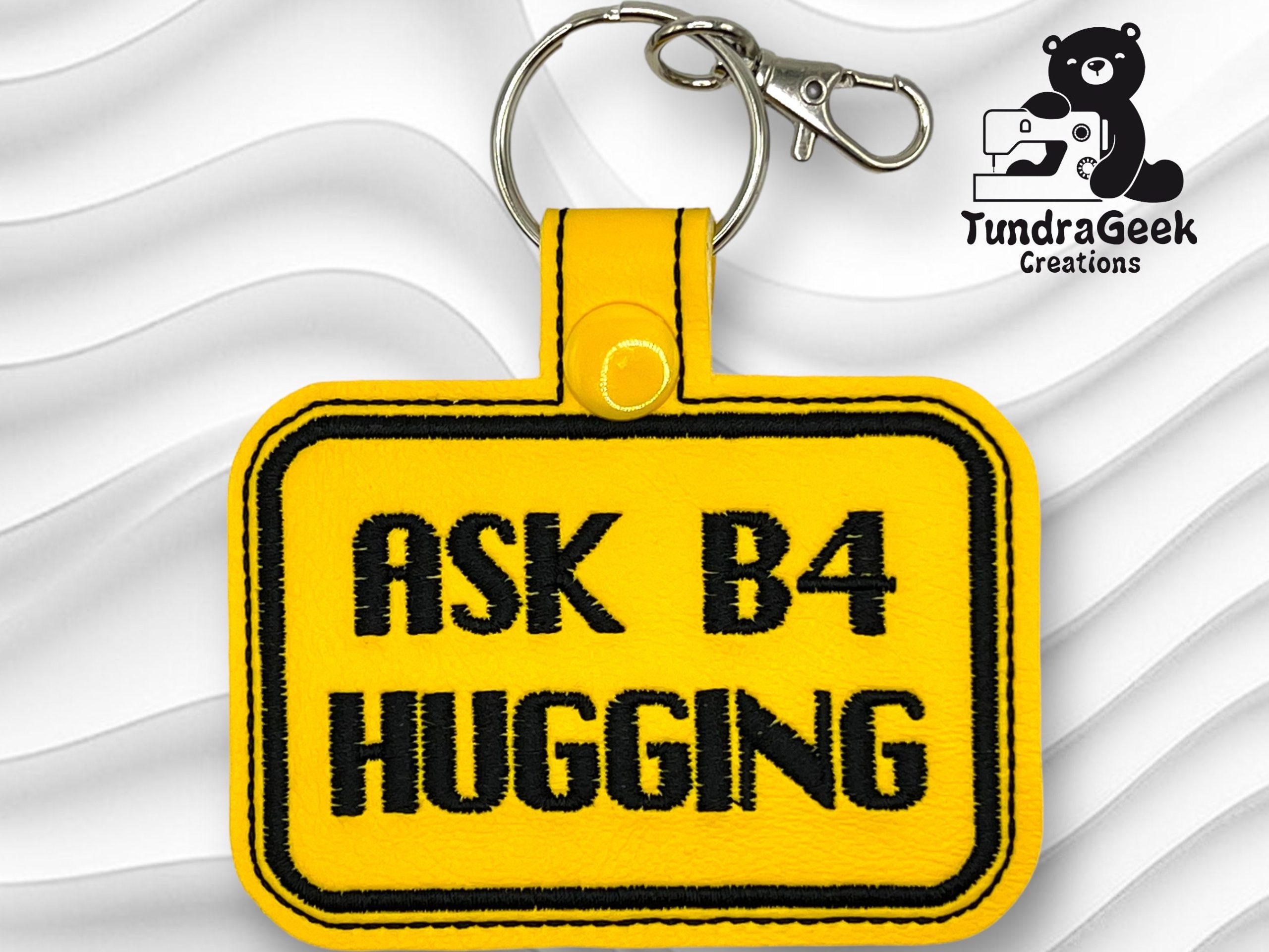 A yellow keychain with black text reads ASK B4 HUGGING. A metal keyring and clasp are attached. The TundraGeek Creations logo is in the top right corner.
