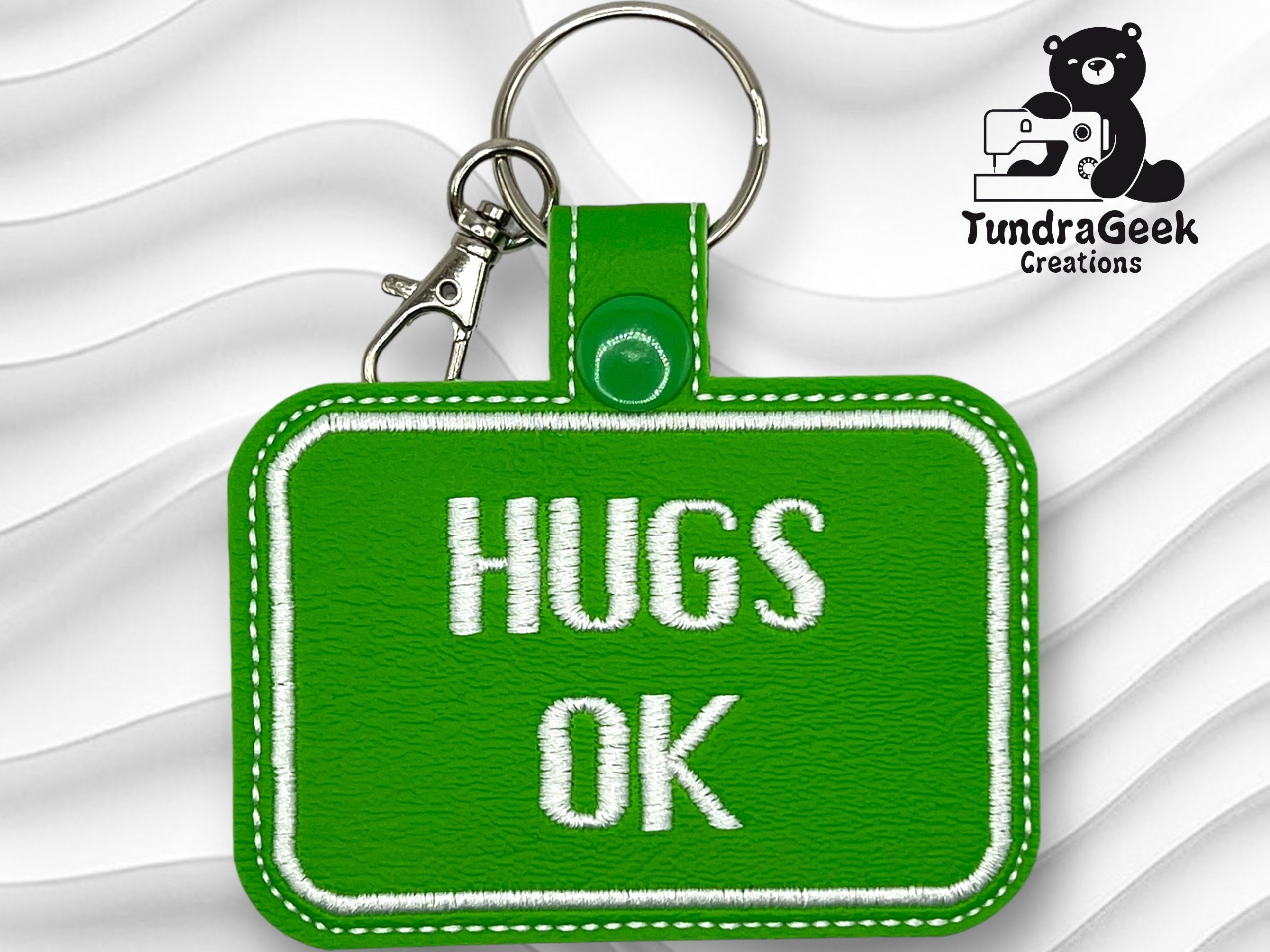 A green keychain with white stitching reads HUGS OK in white letters. It has a metal key ring and a small clasp. The TundraGeek Creations logo is in the top right corner.