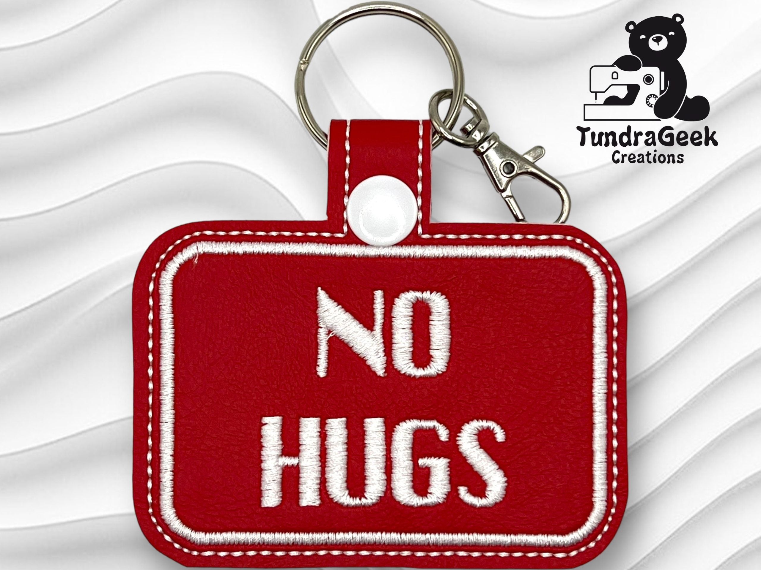 A red rectangular keychain with white stitching and bold white text reading NO HUGS, displayed on a wavy white background with a TundraGeek Creations logo in the corner.